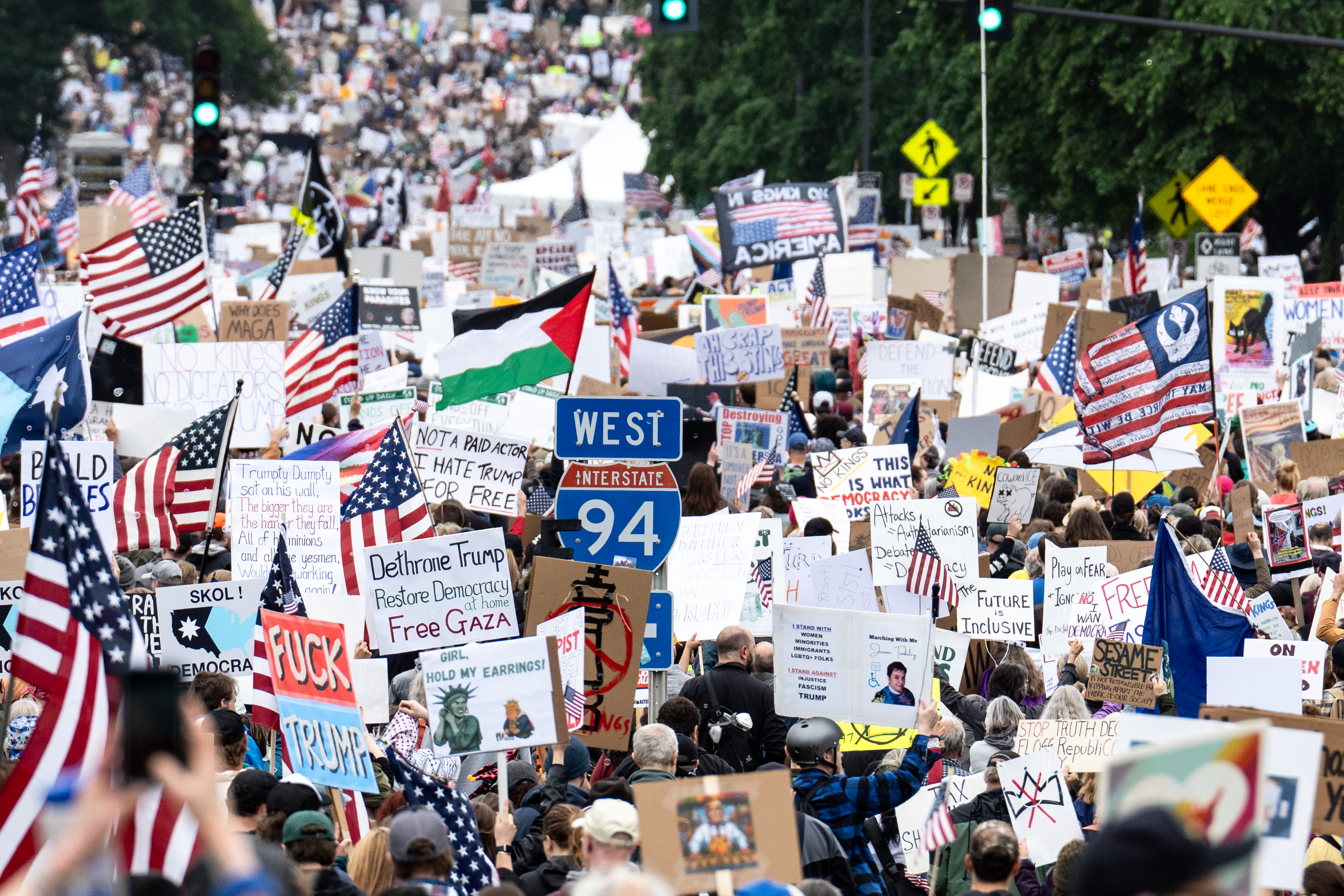 A large crowd fills a street carrying American flags and handmade protest signs at an outdoor rally.