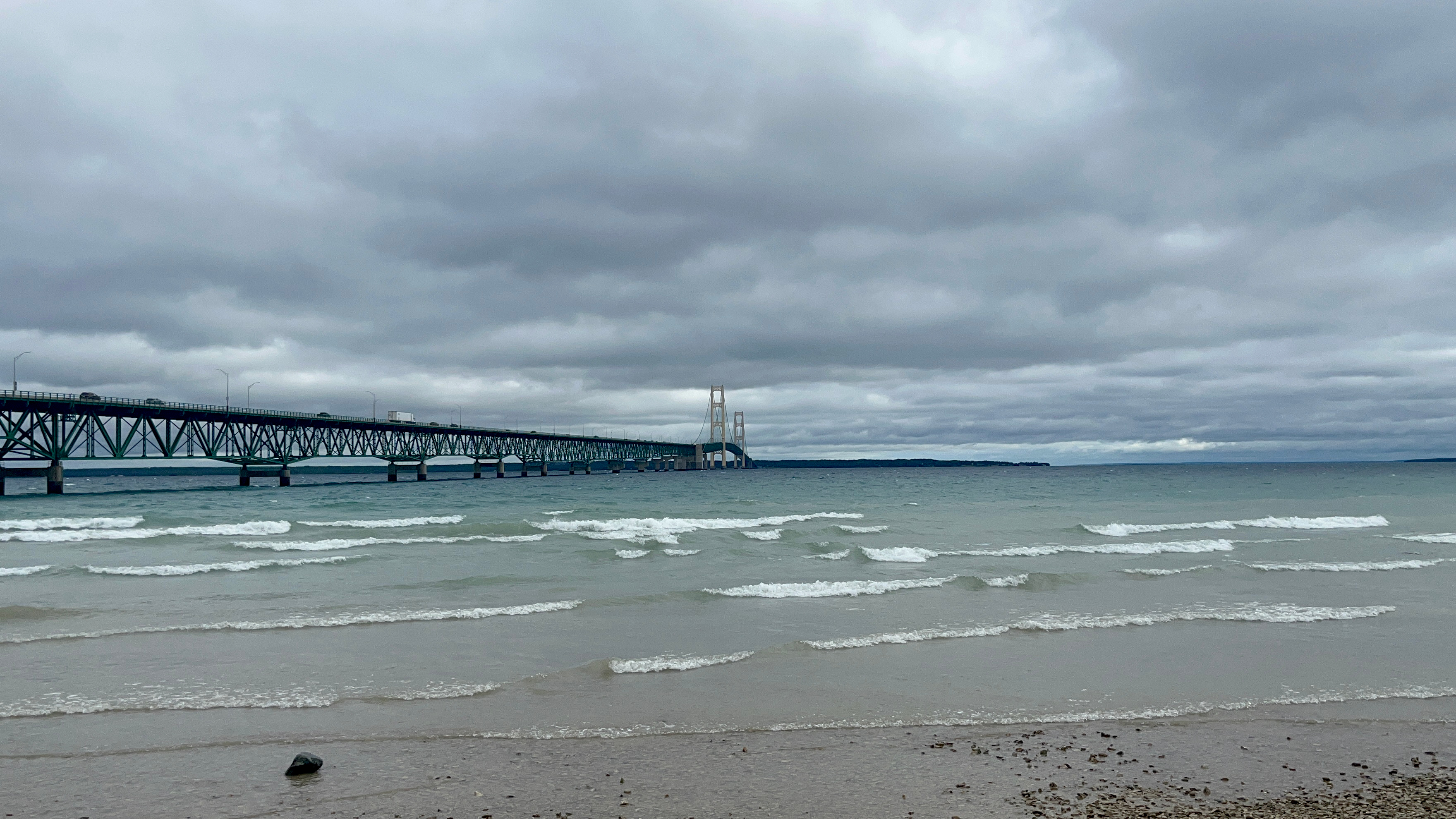 A suspension bridge spans a wide strait, seen from a sandy beach under overcast skies.