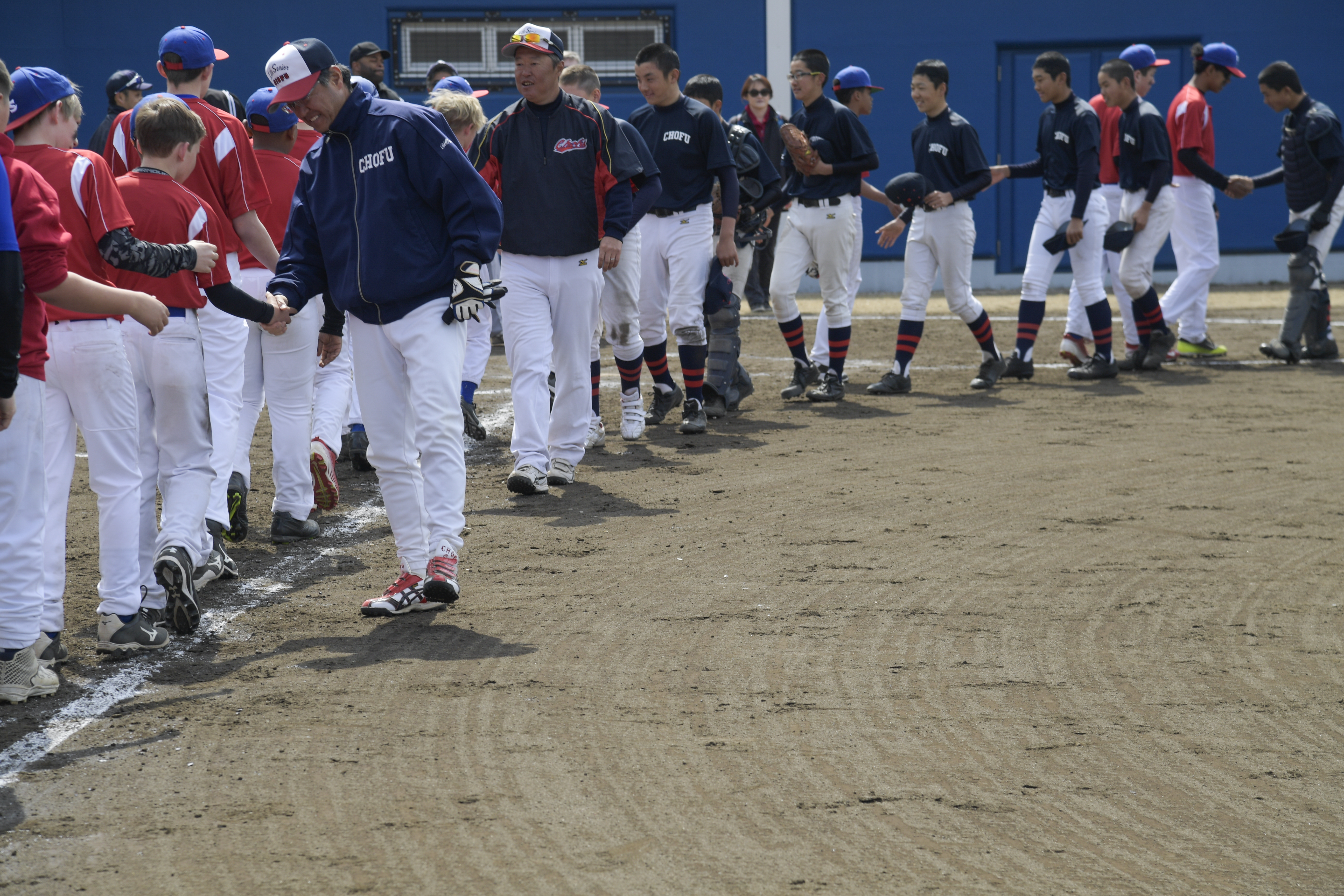 Two lines of youth baseball players in uniforms shaking hands on a dirt field.