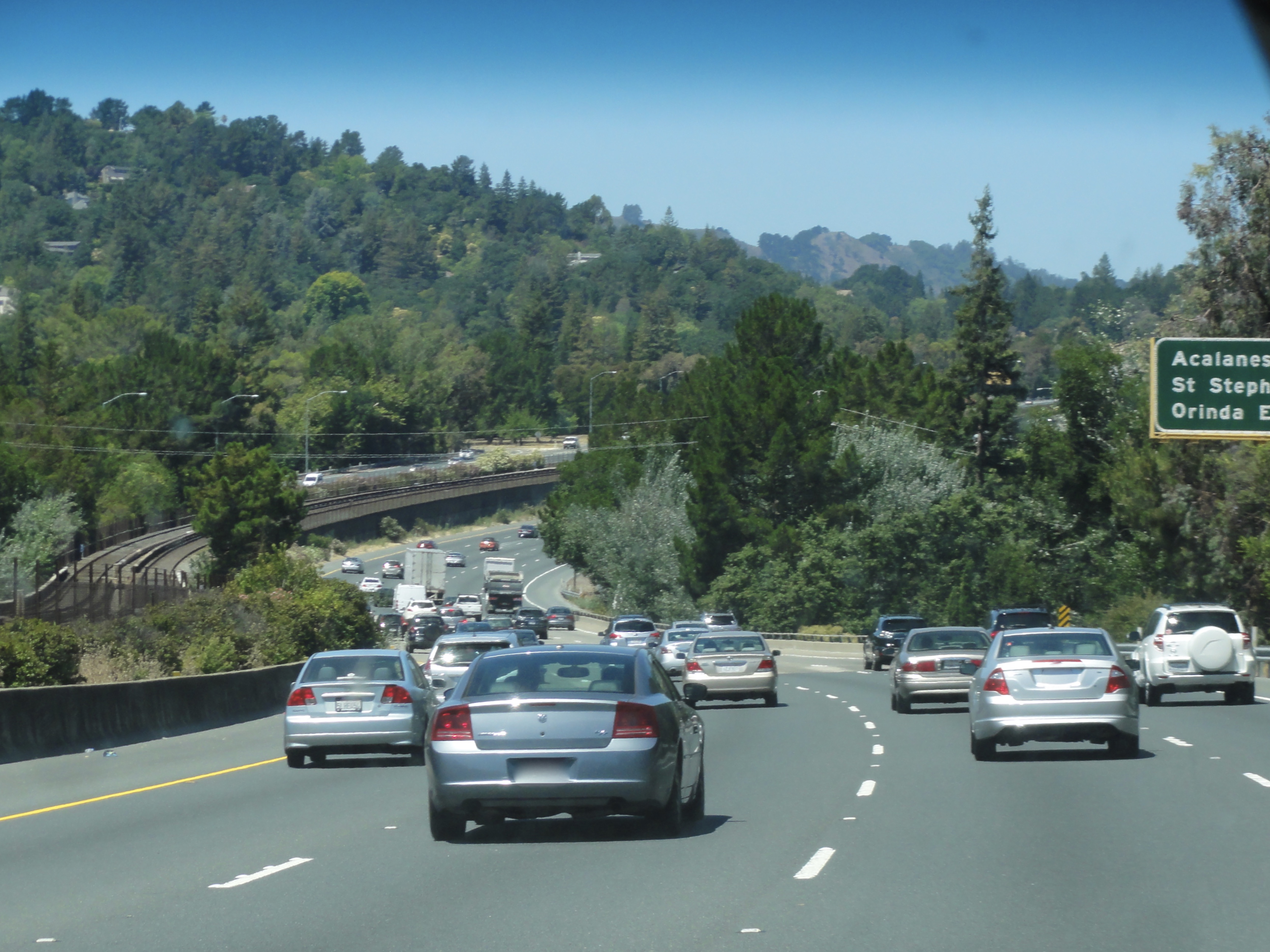 Multiple lanes of eastbound freeway traffic on CA Route 24 with tree-covered hills on both sides.