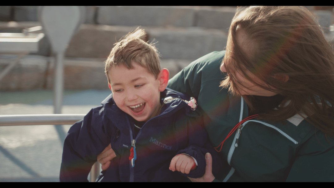 A young boy laughing joyfully while being held by a woman outdoors, both wearing jackets.