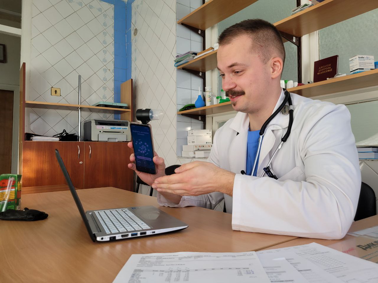 A clinician wearing a white coat and stethoscope holds a smartphone beside an open laptop at a desk in a medical office.