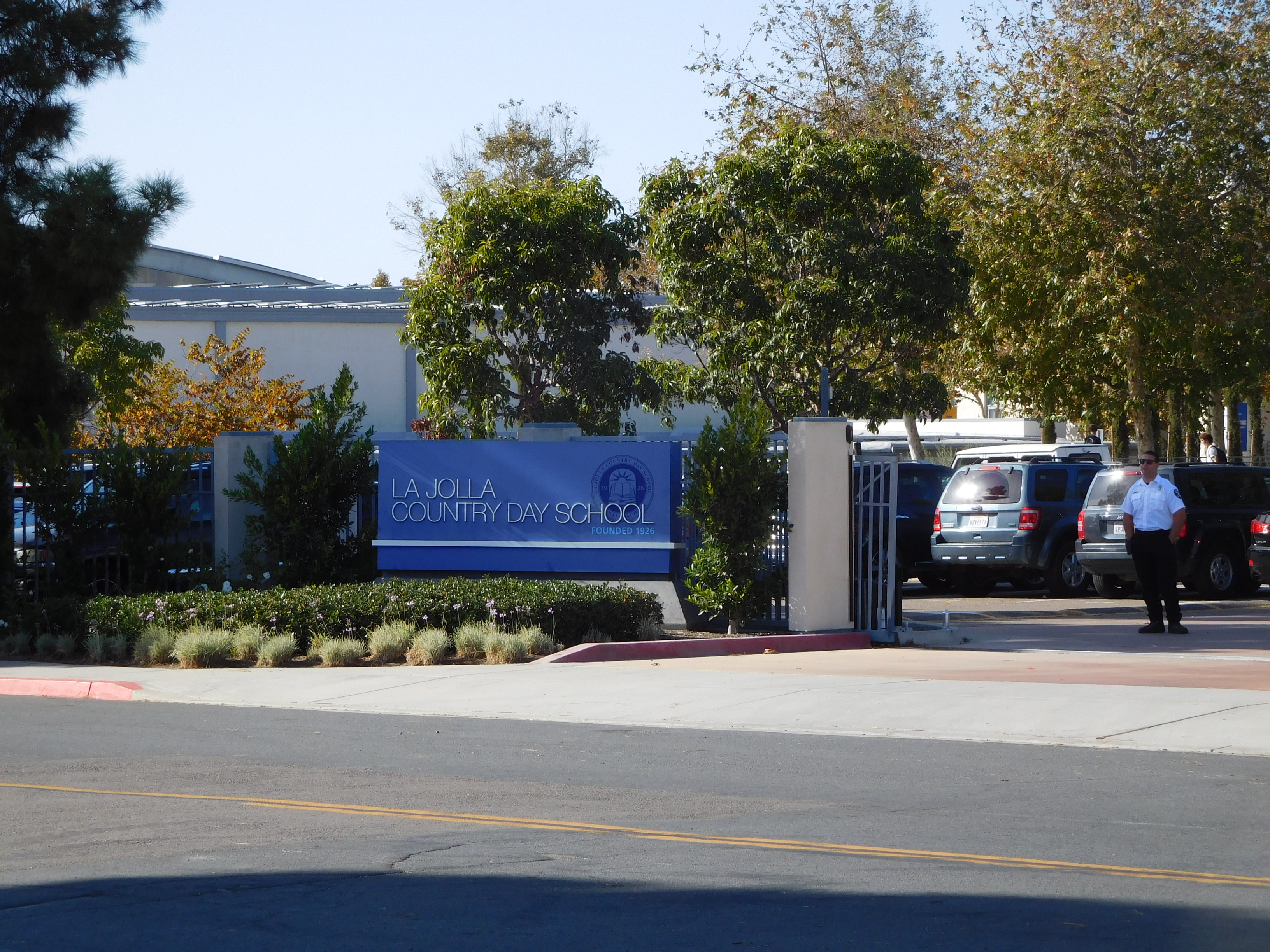 Gated school entrance with a blue sign, mature trees, and campus buildings behind.
