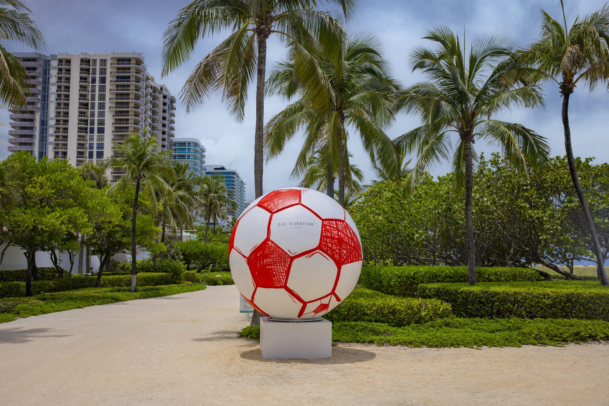 A large red and white soccer ball sculpture on a pedestal surrounded by palm trees and high-rise buildings.