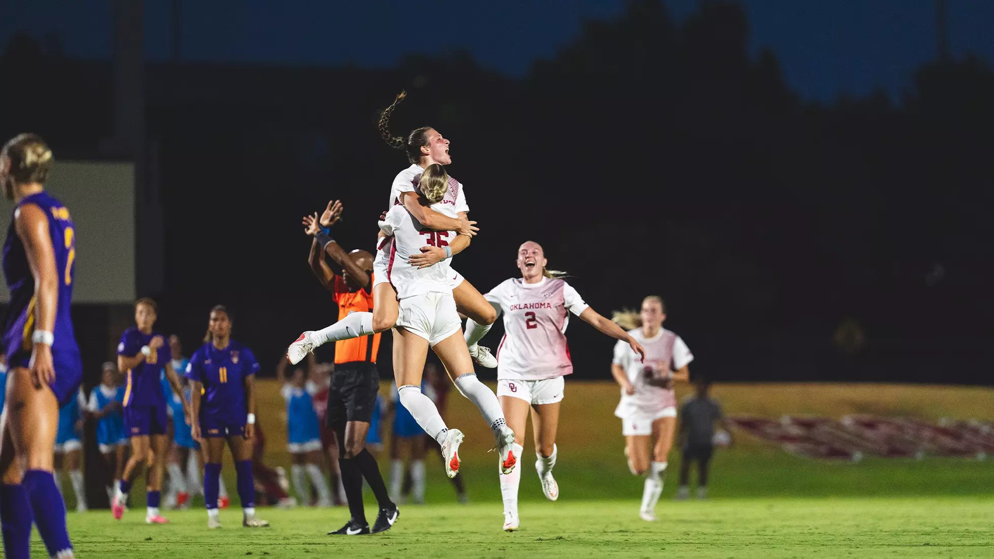 Two soccer players in white uniforms jump and embrace in celebration on a lit field at night.