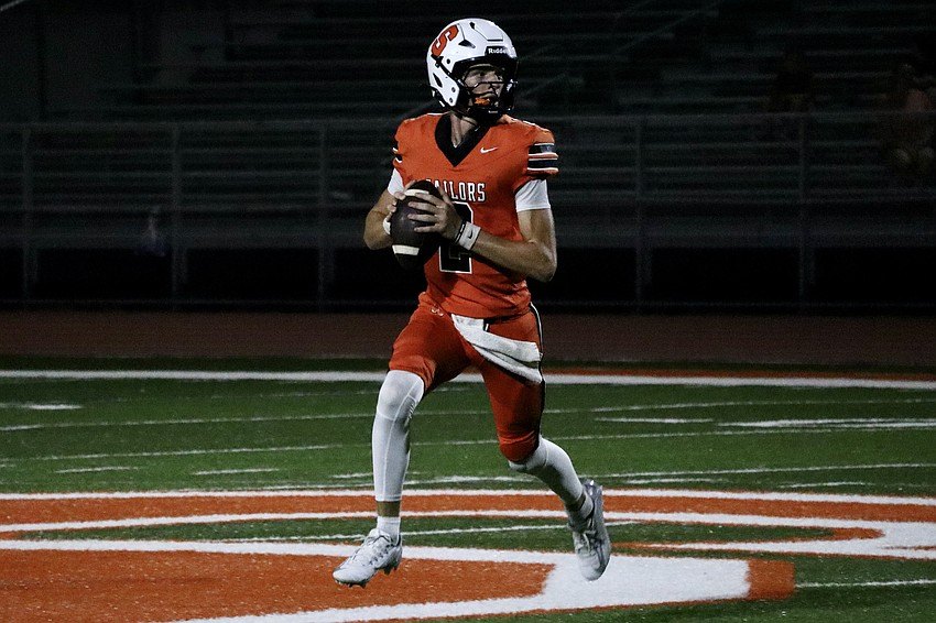 A football player in an orange jersey holds a football in a passing stance on a lit field at night.