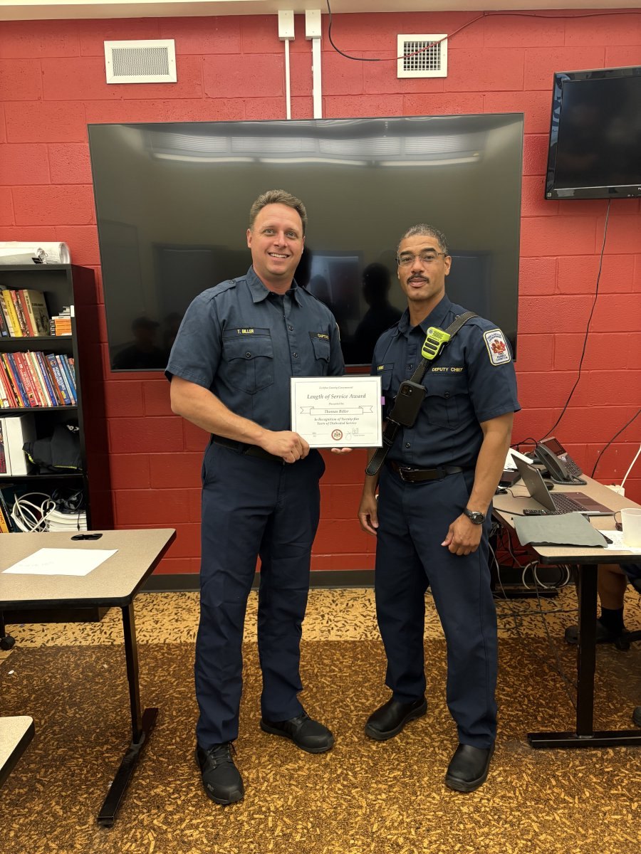 Two uniformed fire department personnel holding a certificate in an indoor meeting room.