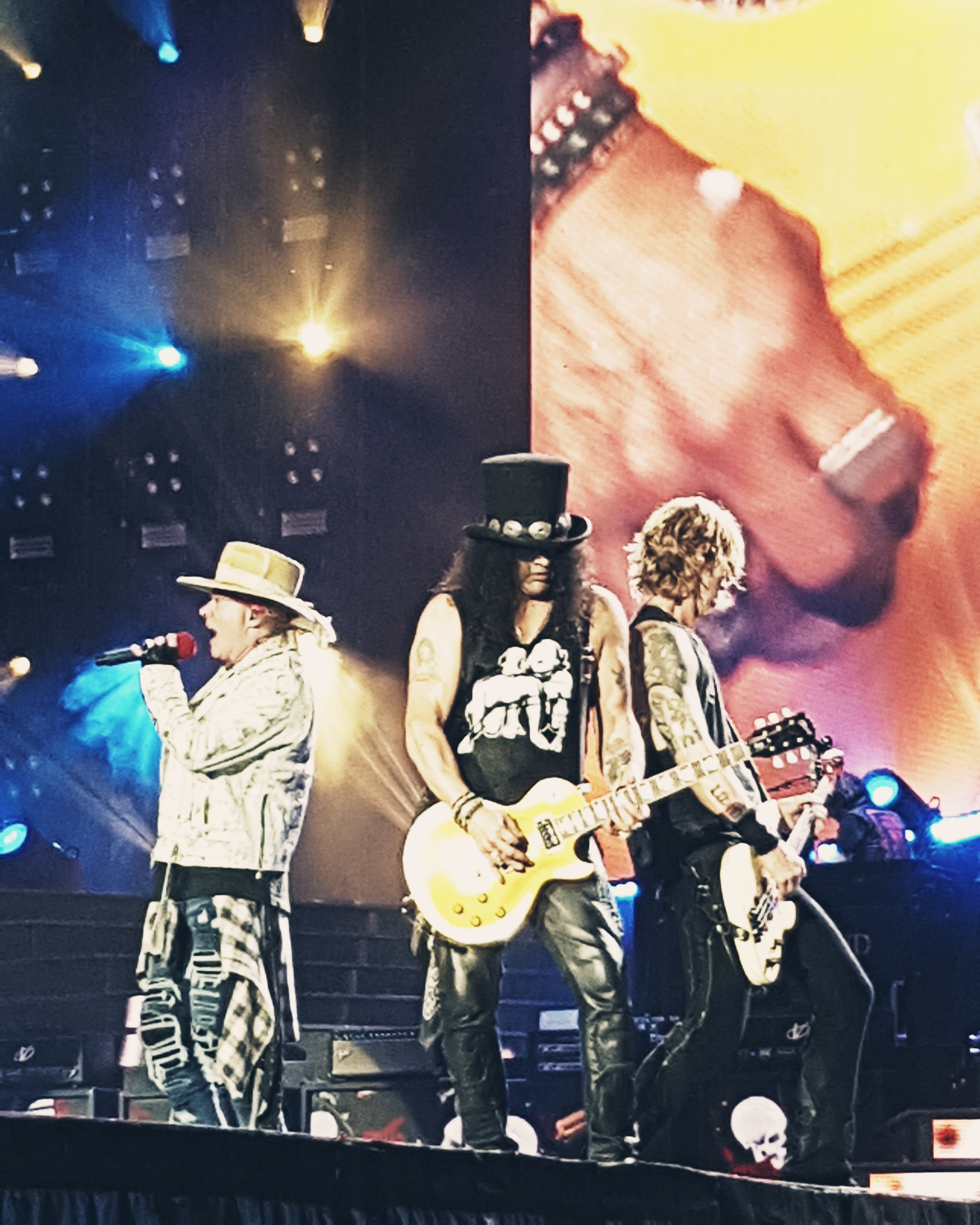 Three rock musicians performing on a large lit concert stage with a video screen backdrop.