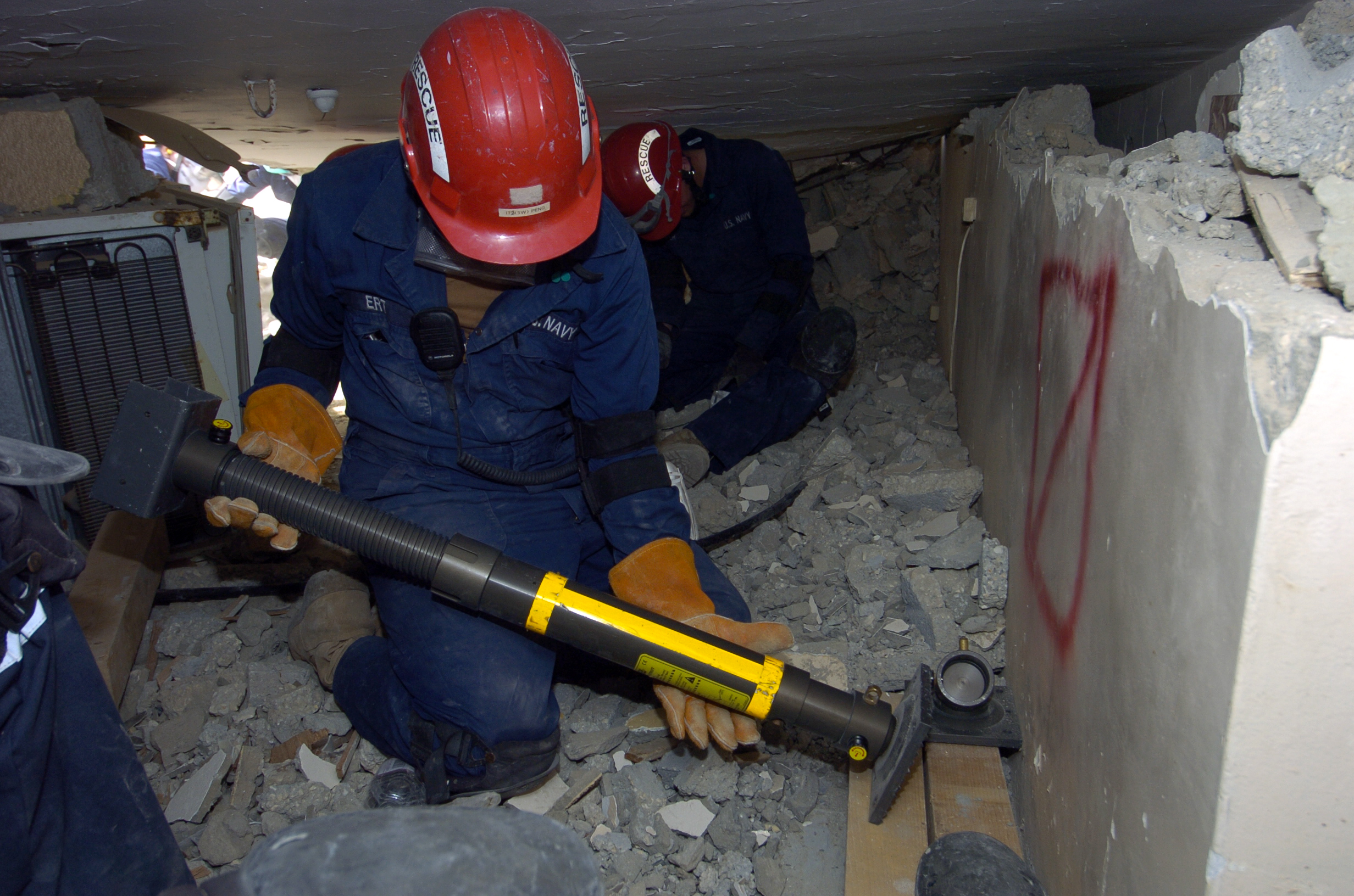 Two workers in hard hats brace a hydraulic strut inside a rubble-filled collapsed structure.
