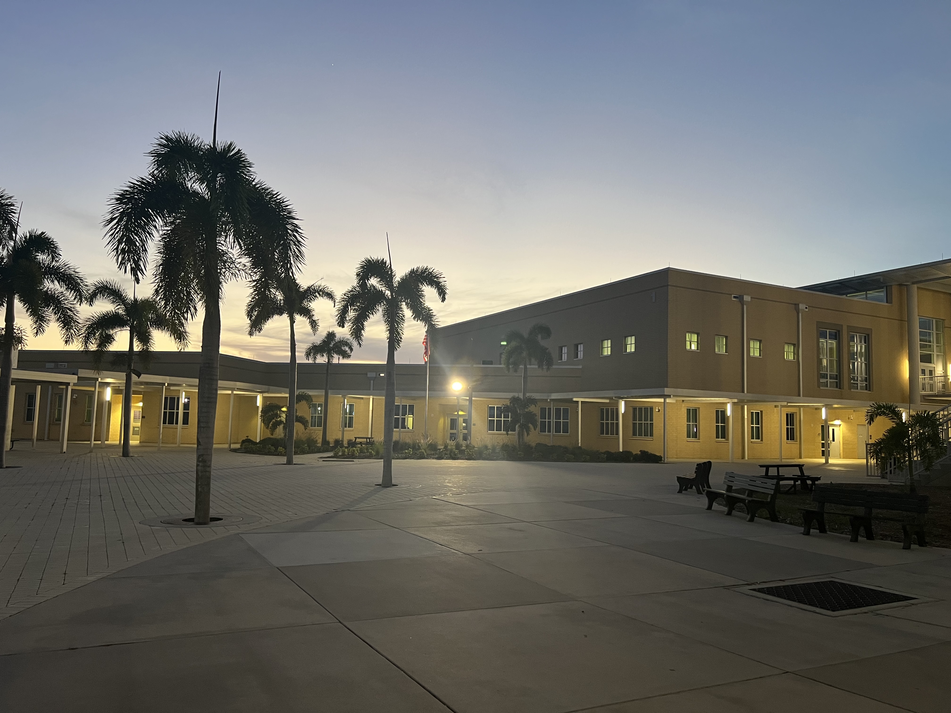 School campus courtyard at dusk with a large yellow two-story building and palm trees.