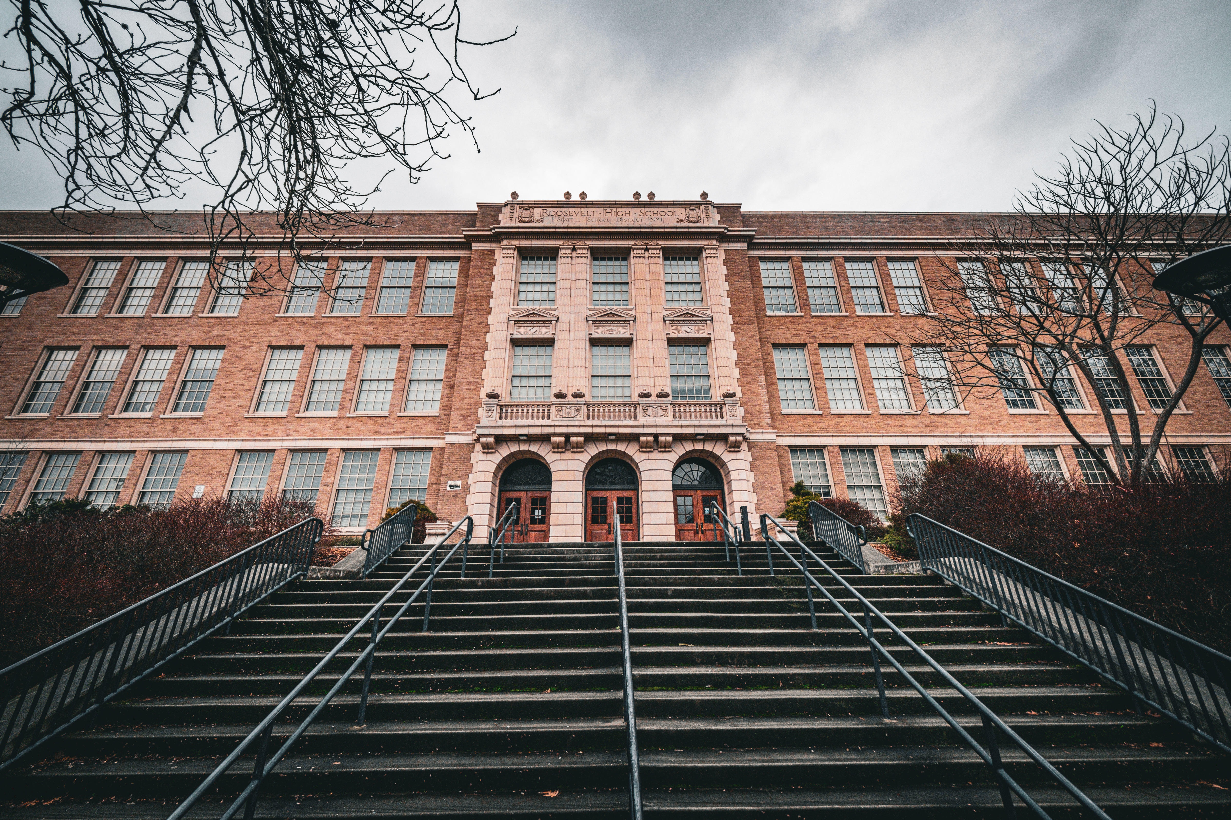 Red-brick high school building viewed from the bottom of a wide staircase under an overcast sky.