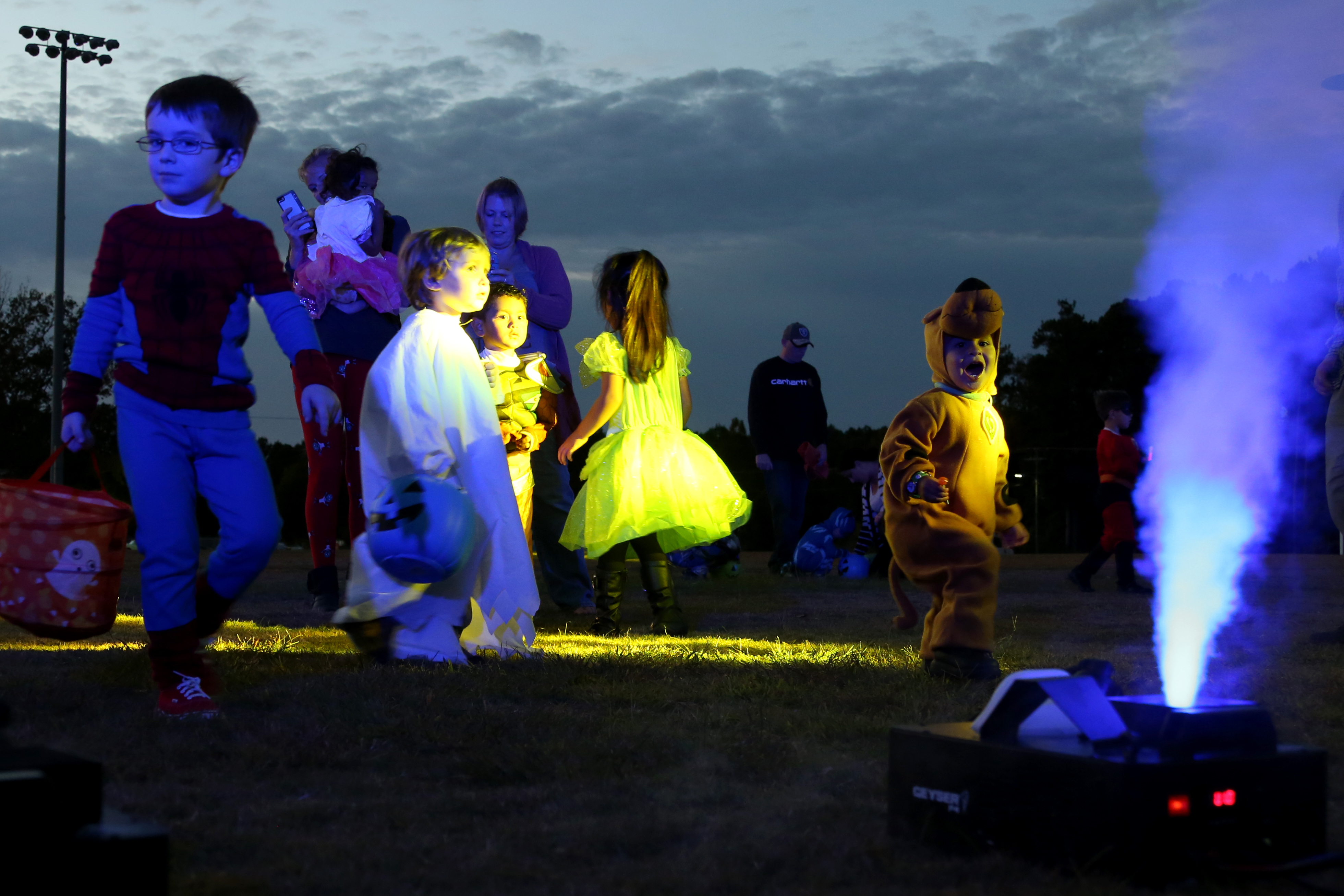 Children in Halloween costumes play outdoors at dusk near a glowing fog machine.