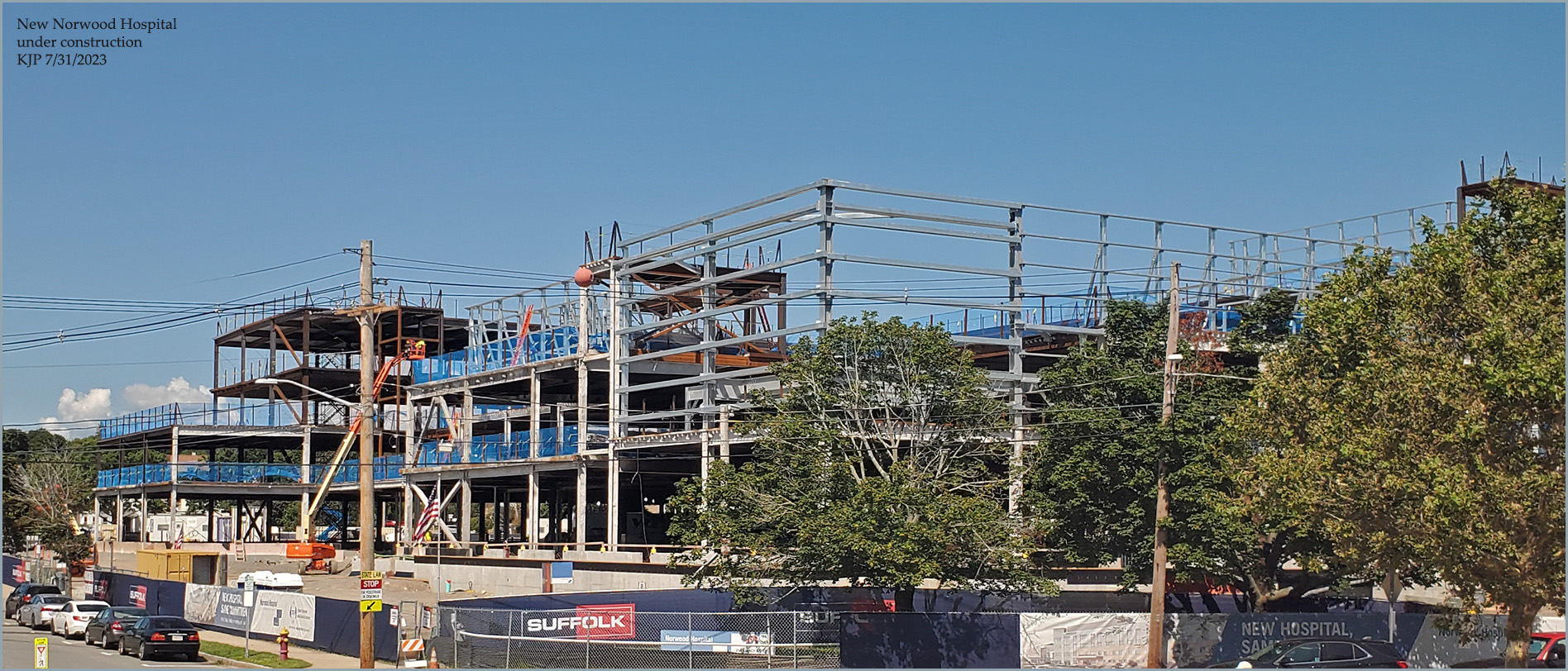 Multi-story steel frame of a hospital building under construction with cranes and contractor fencing.