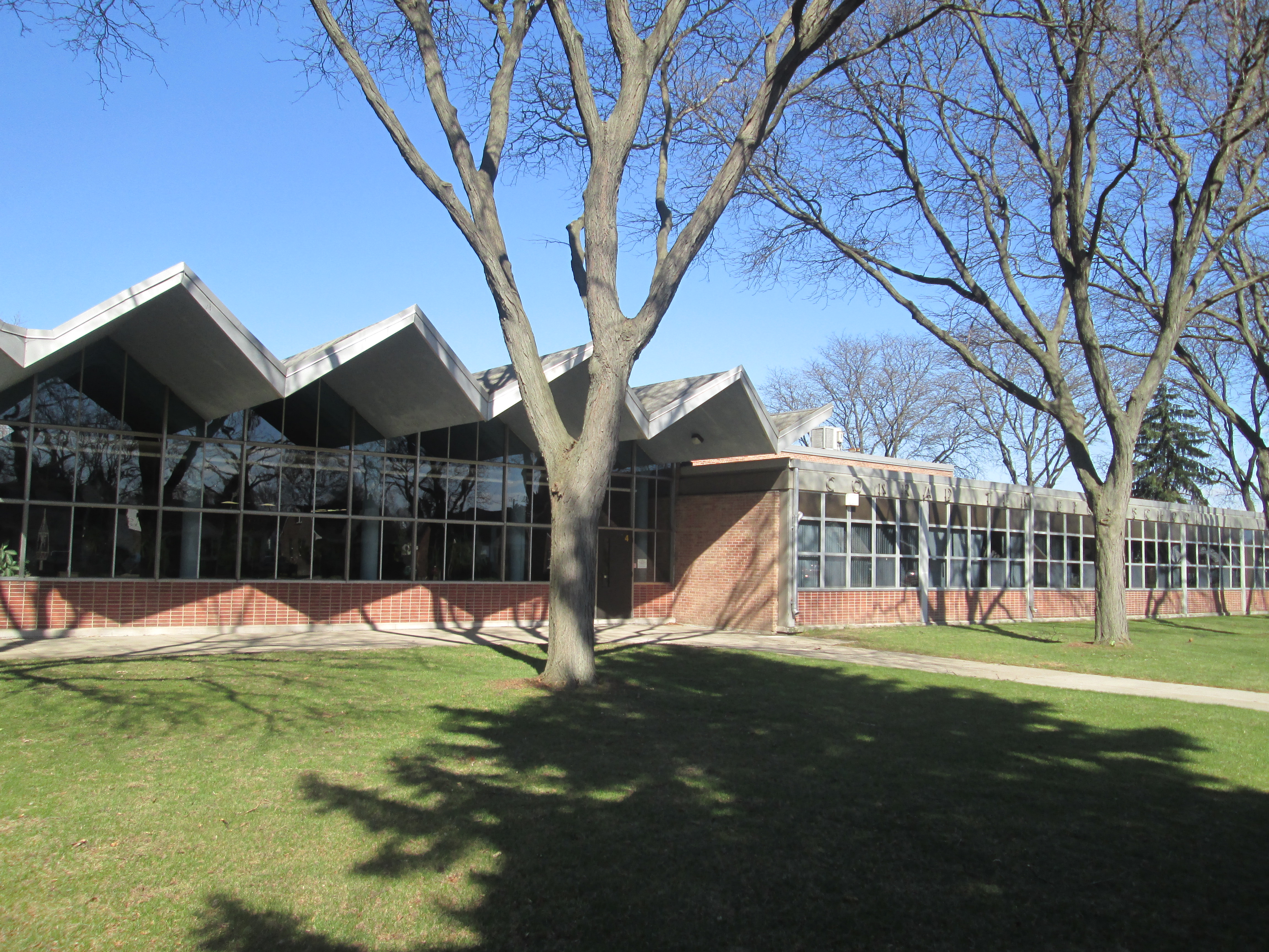 Mid-century brick and glass school administration building with a sawtooth roofline and bare trees