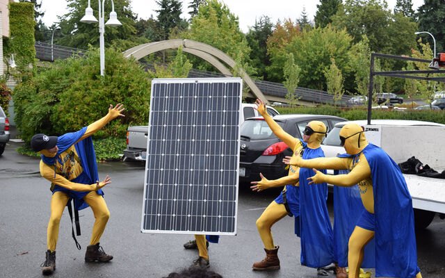 Three people in yellow bodysuits and blue capes hold a solar panel in a parking lot.