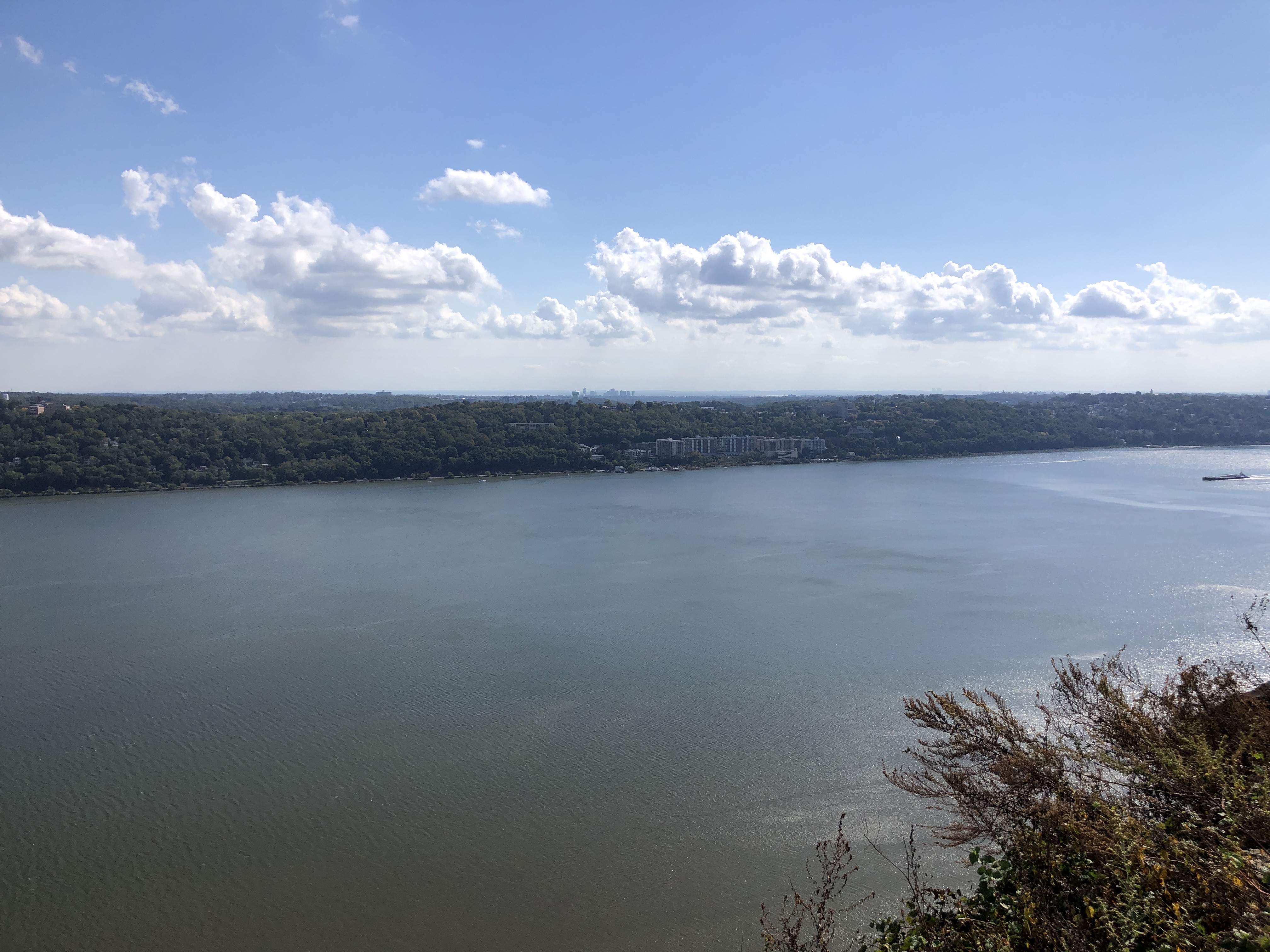 Wide view of the Hudson River with a barge and tree-lined shores under partly cloudy skies.