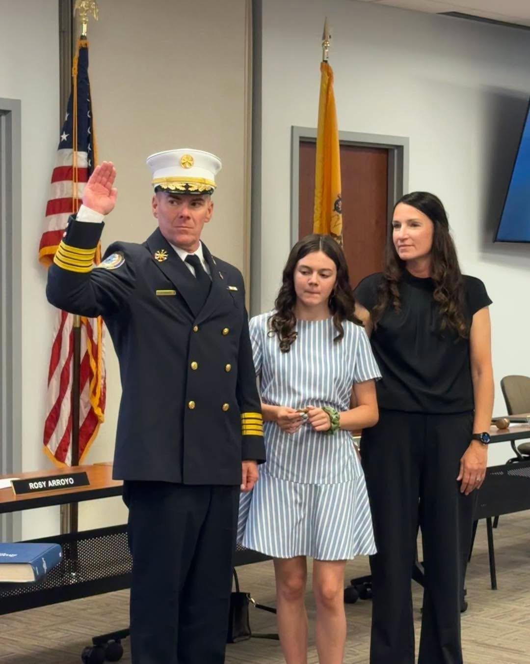 Man in white fire chief dress uniform raises right hand taking an oath, flanked by two women.