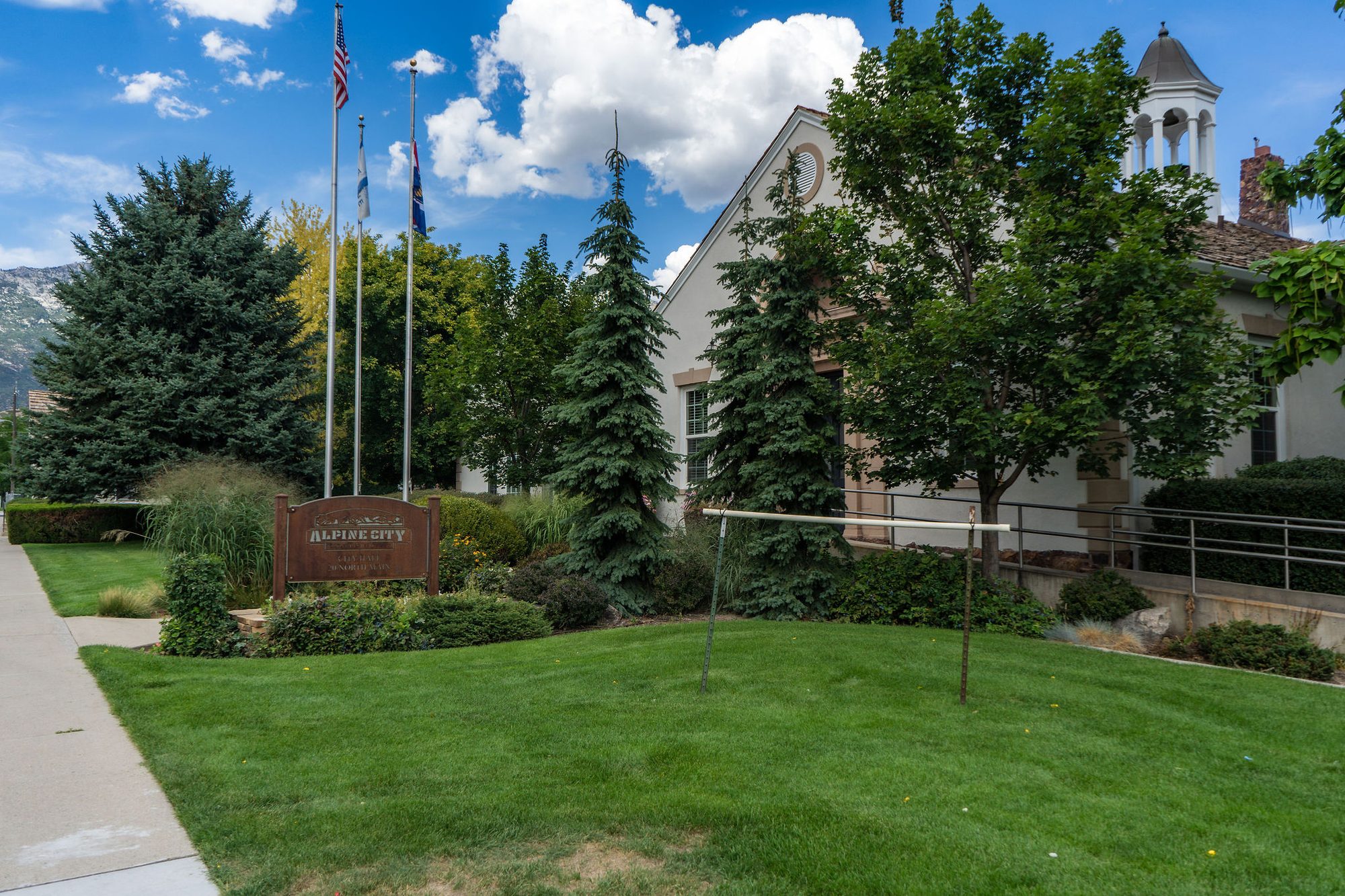 Civic building with bell tower behind a manicured lawn and three flagpoles, mountains in background.