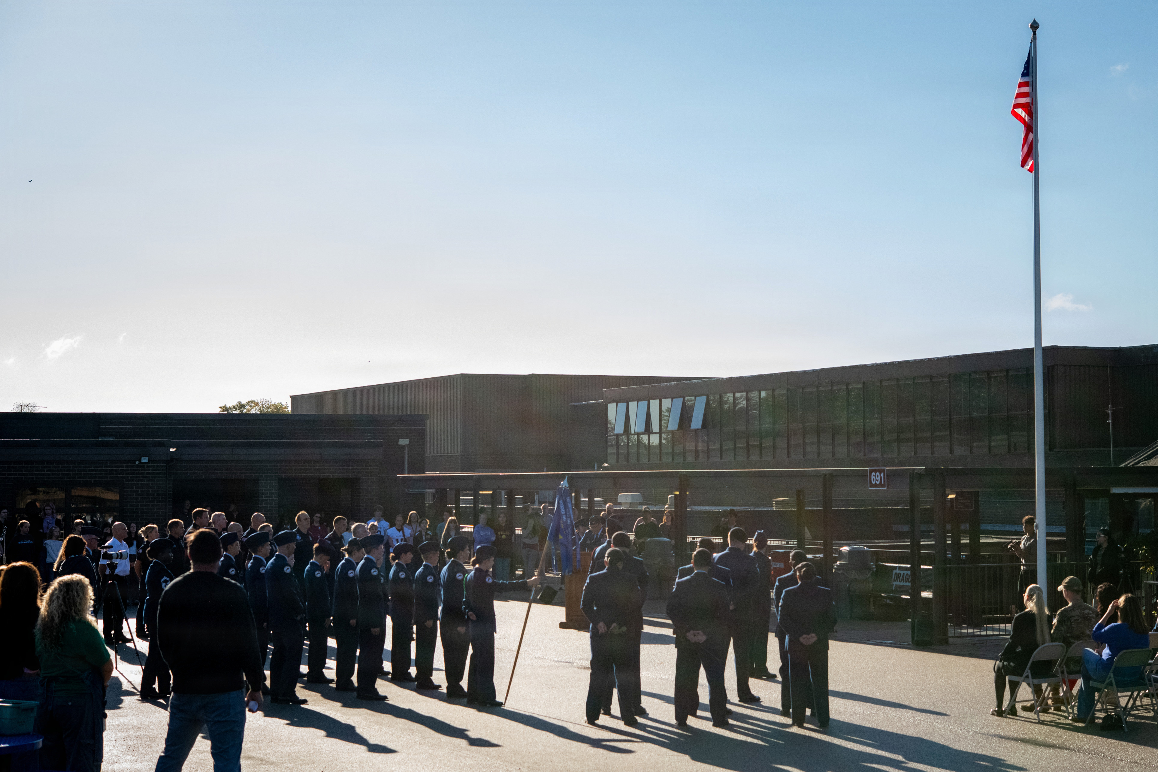Uniformed personnel and civilians gathered outdoors at a 9/11 anniversary memorial ceremony under an American flag.