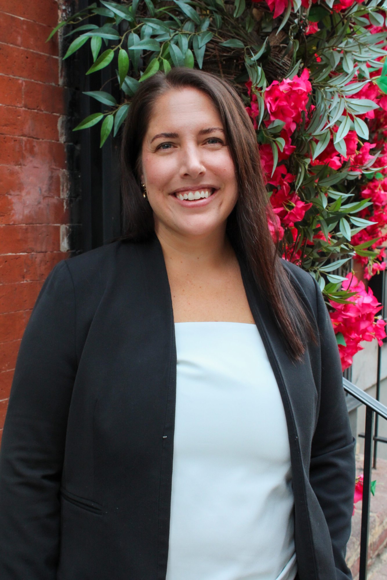 Woman with long dark hair in a black blazer smiling outdoors in front of a brick wall.