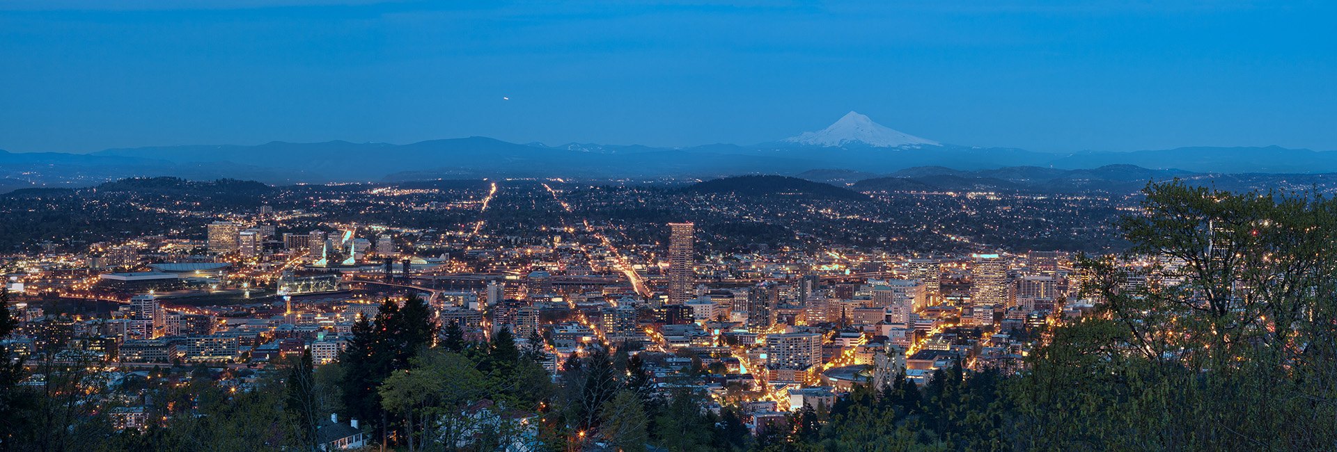 Panoramic dusk aerial view of a city skyline with illuminated streets and a snow-capped mountain on the horizon.