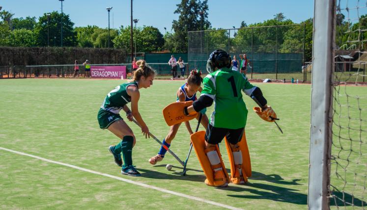 Female field hockey players and a goalkeeper compete near a goal on a turf field.