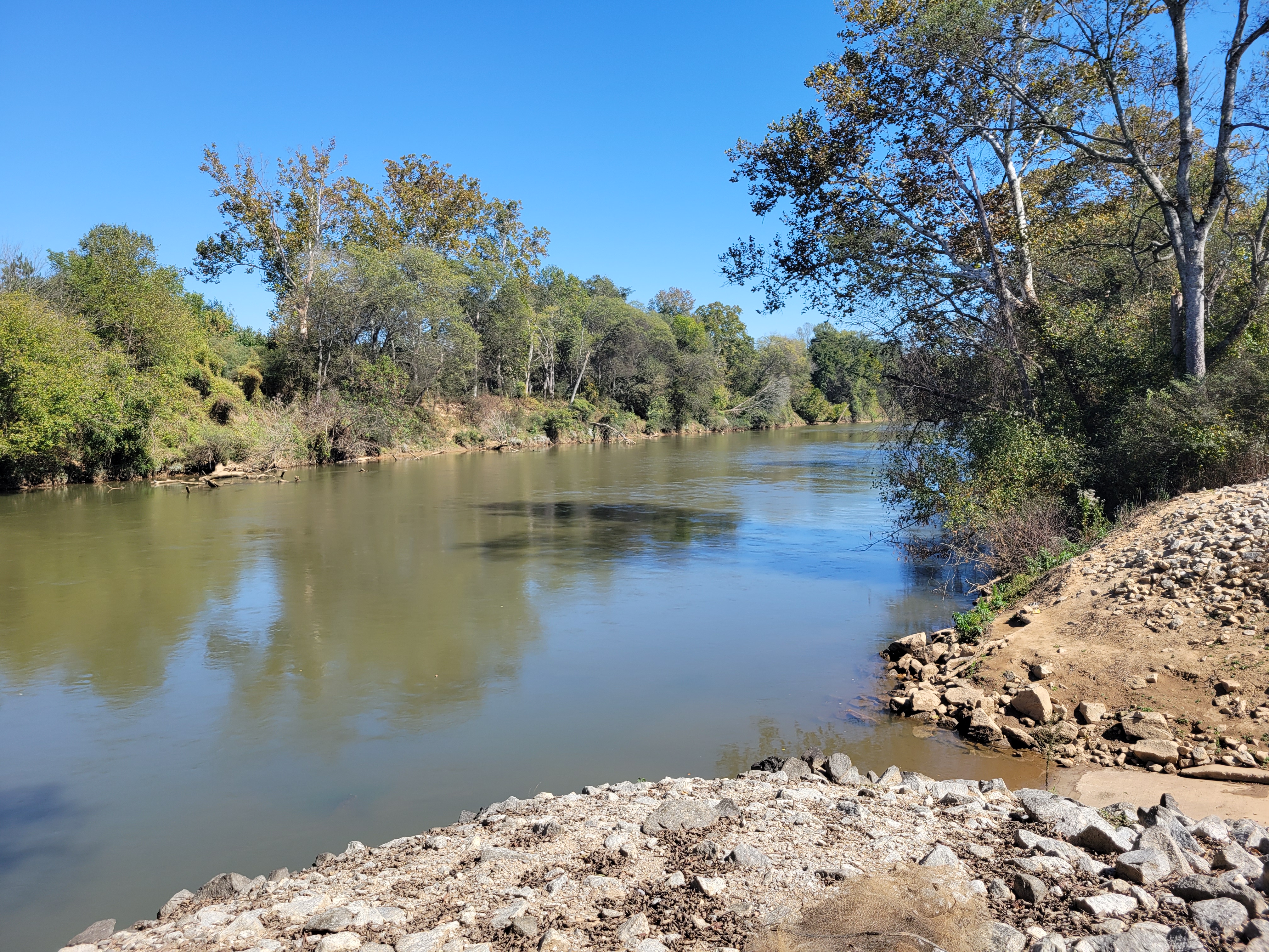 Calm river with rocky shoreline and dense tree-lined banks under a blue sky.