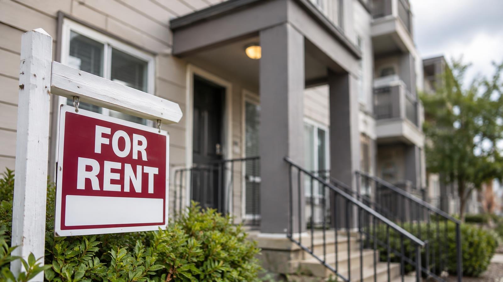 Small multi-unit apartment building with a for-rent sign near the entrance on a suburban street.