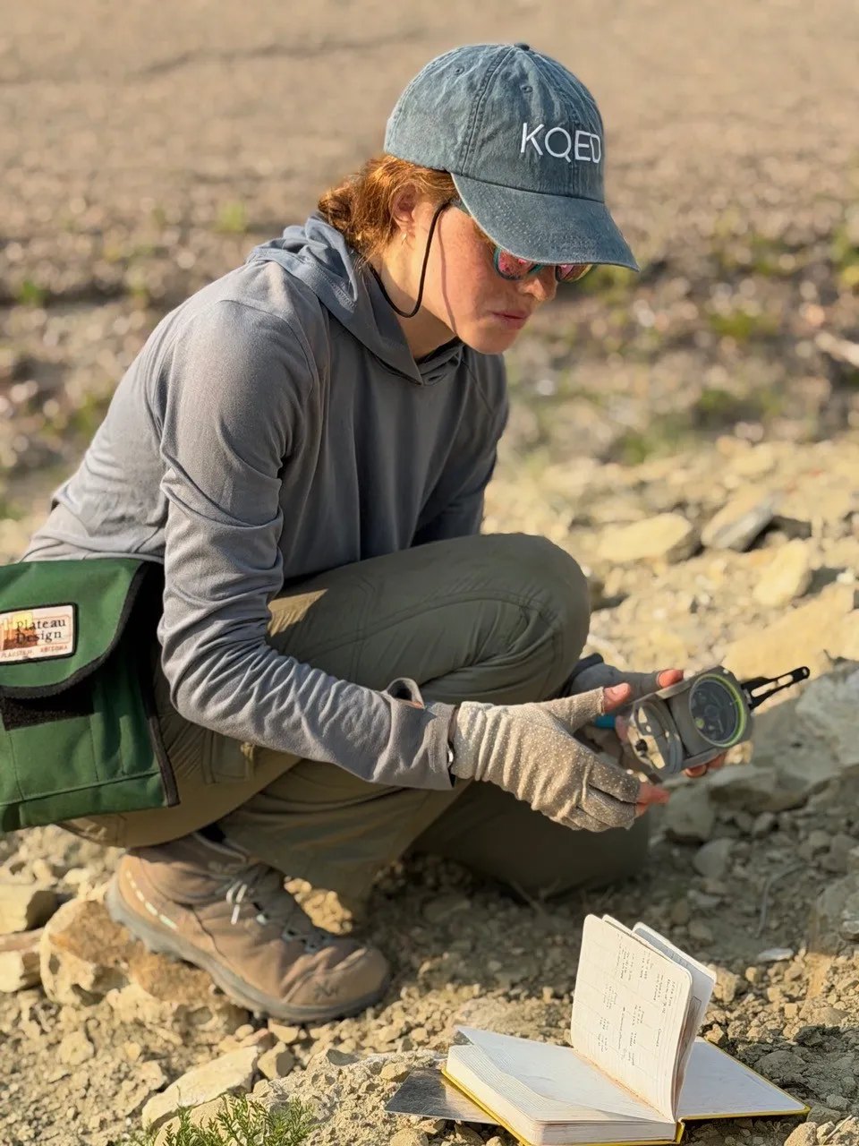 Student crouching on rocky desert terrain, holding a measuring instrument over an open field notebook.