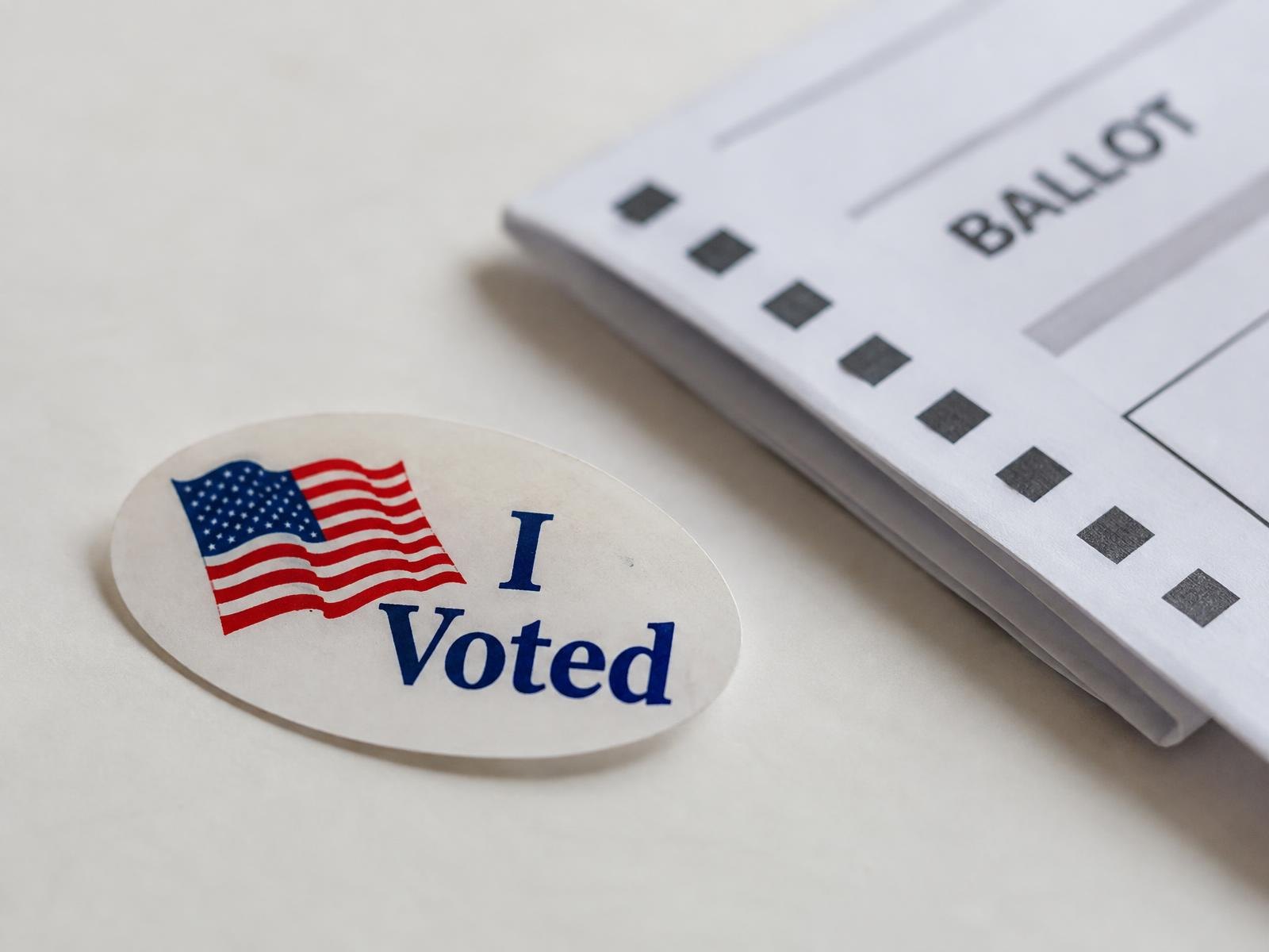 An I Voted sticker resting on a white surface next to a folded paper ballot.