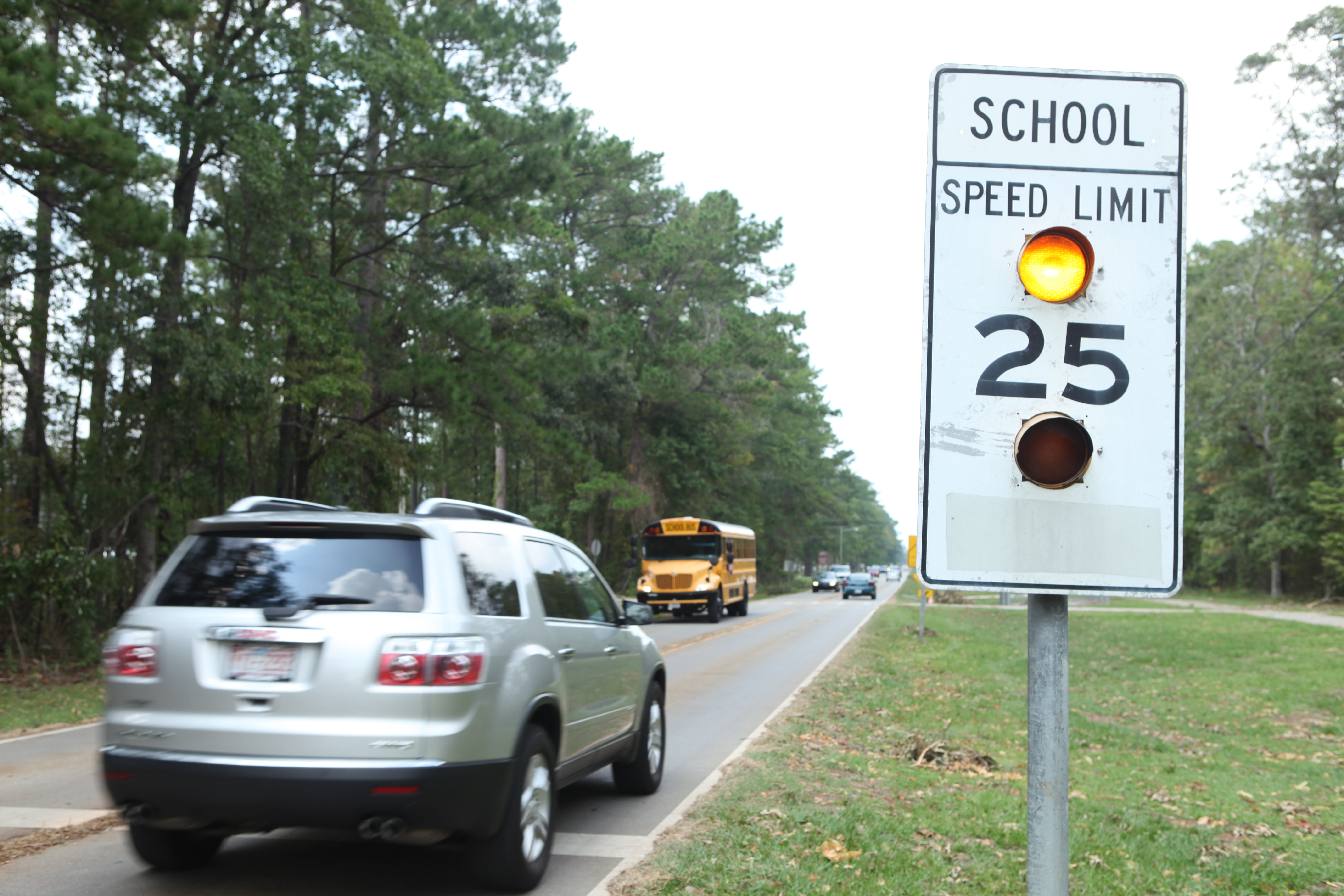 School speed limit 25 mph sign with flashing yellow beacons on a tree-lined road, school bus in background.