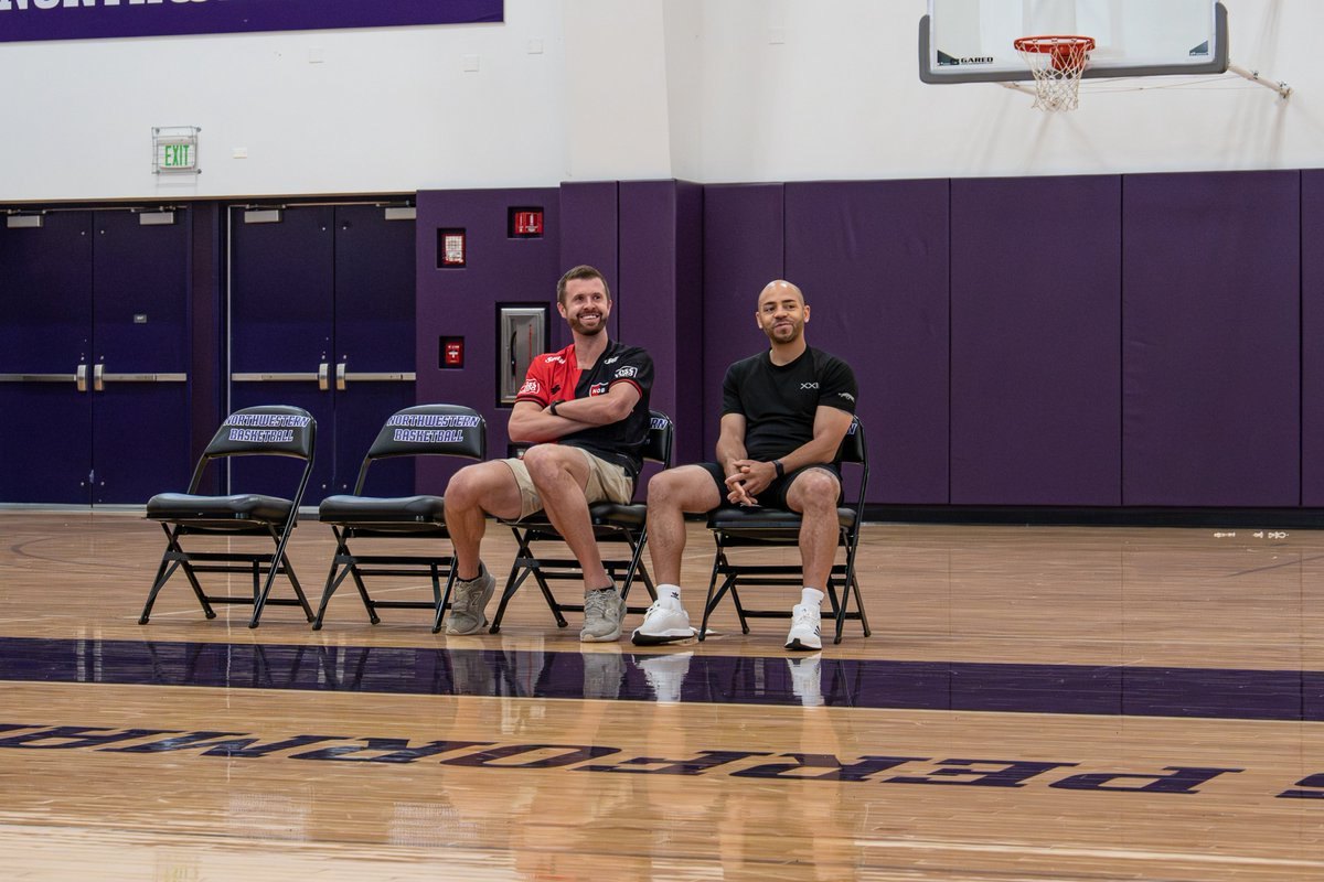 Two men sitting on folding chairs courtside in a basketball gym with purple padded walls.