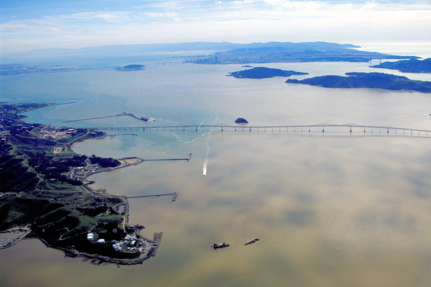 Aerial view of a long bridge spanning a bay with surrounding shoreline and distant mountains.