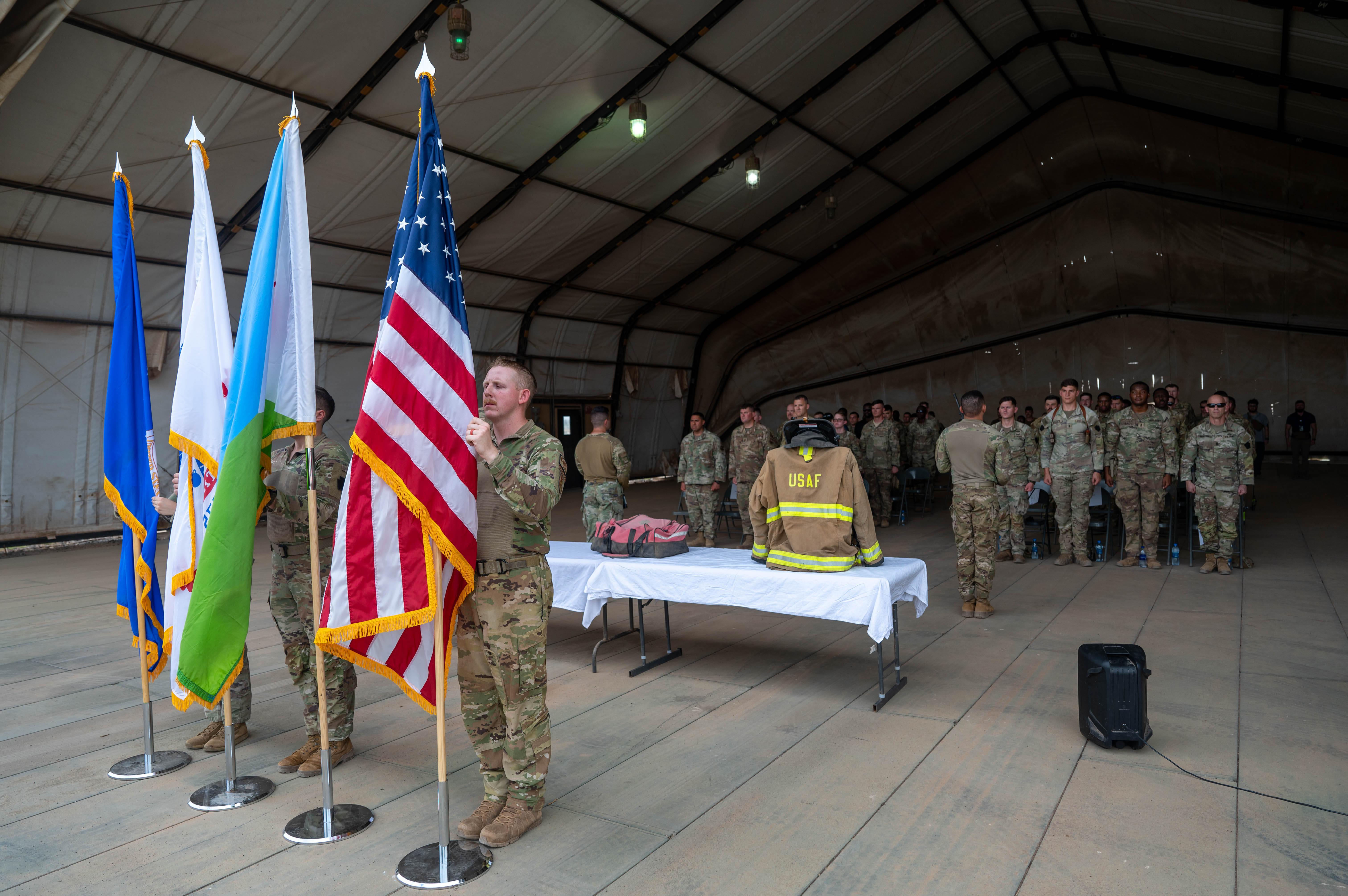 Military color guard posts flags at a 9/11 memorial ceremony, with firefighter gear on a display table.