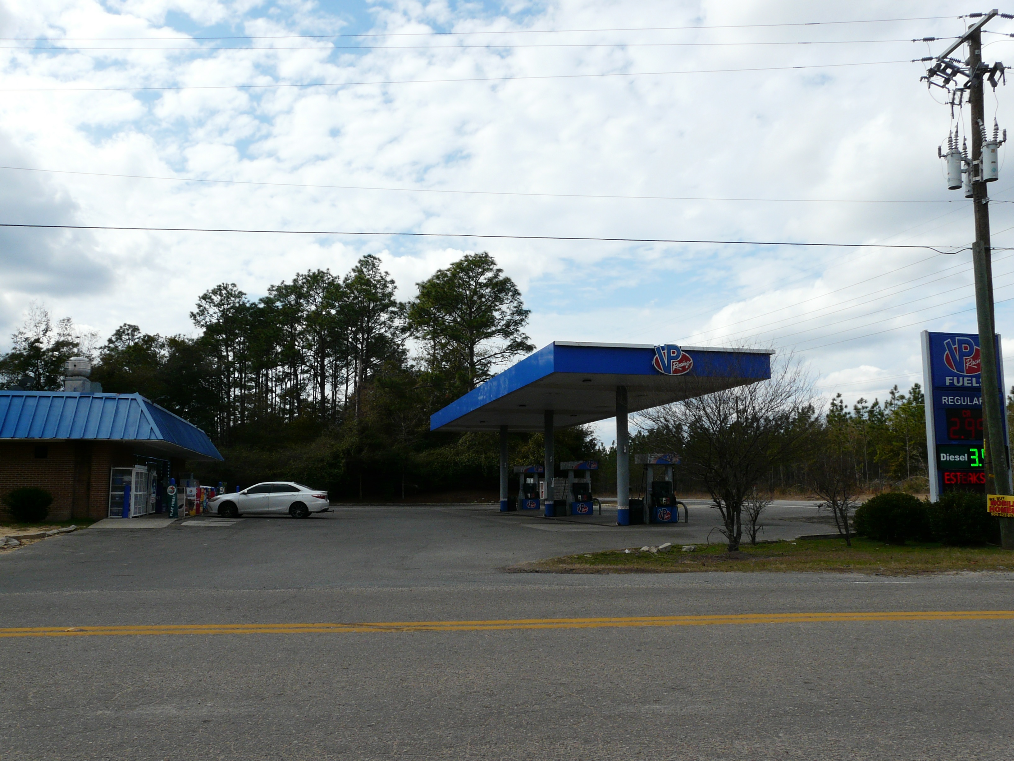 Gas station with blue canopy, fuel pumps, and a digital price sign showing fuel prices.
