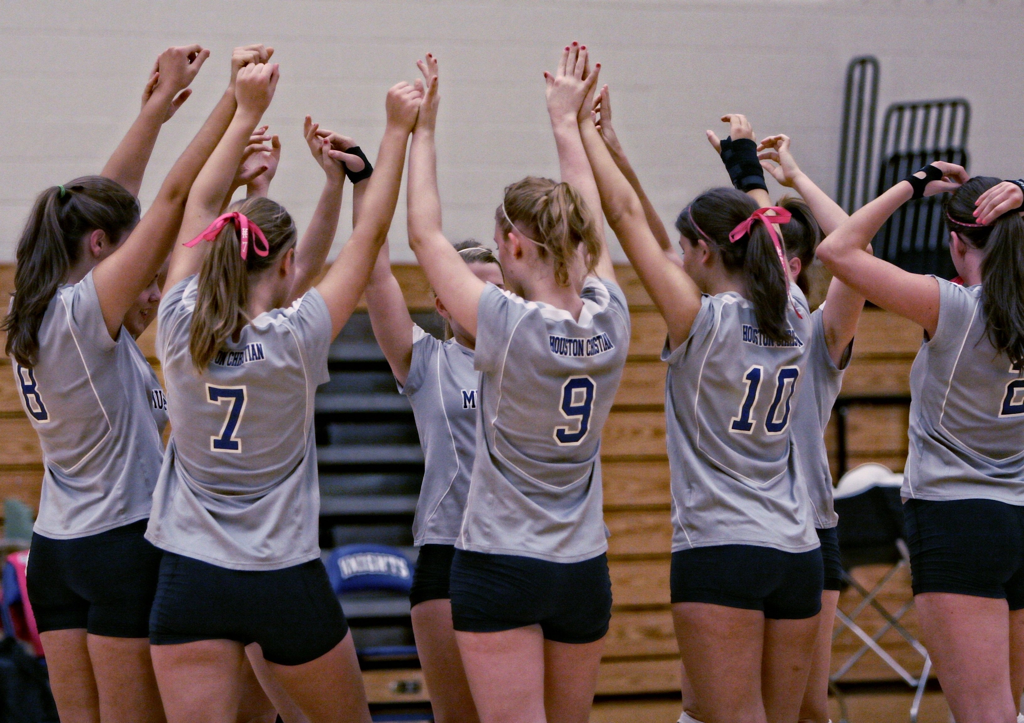 Girls volleyball players in gray uniforms raise arms together in a team huddle.