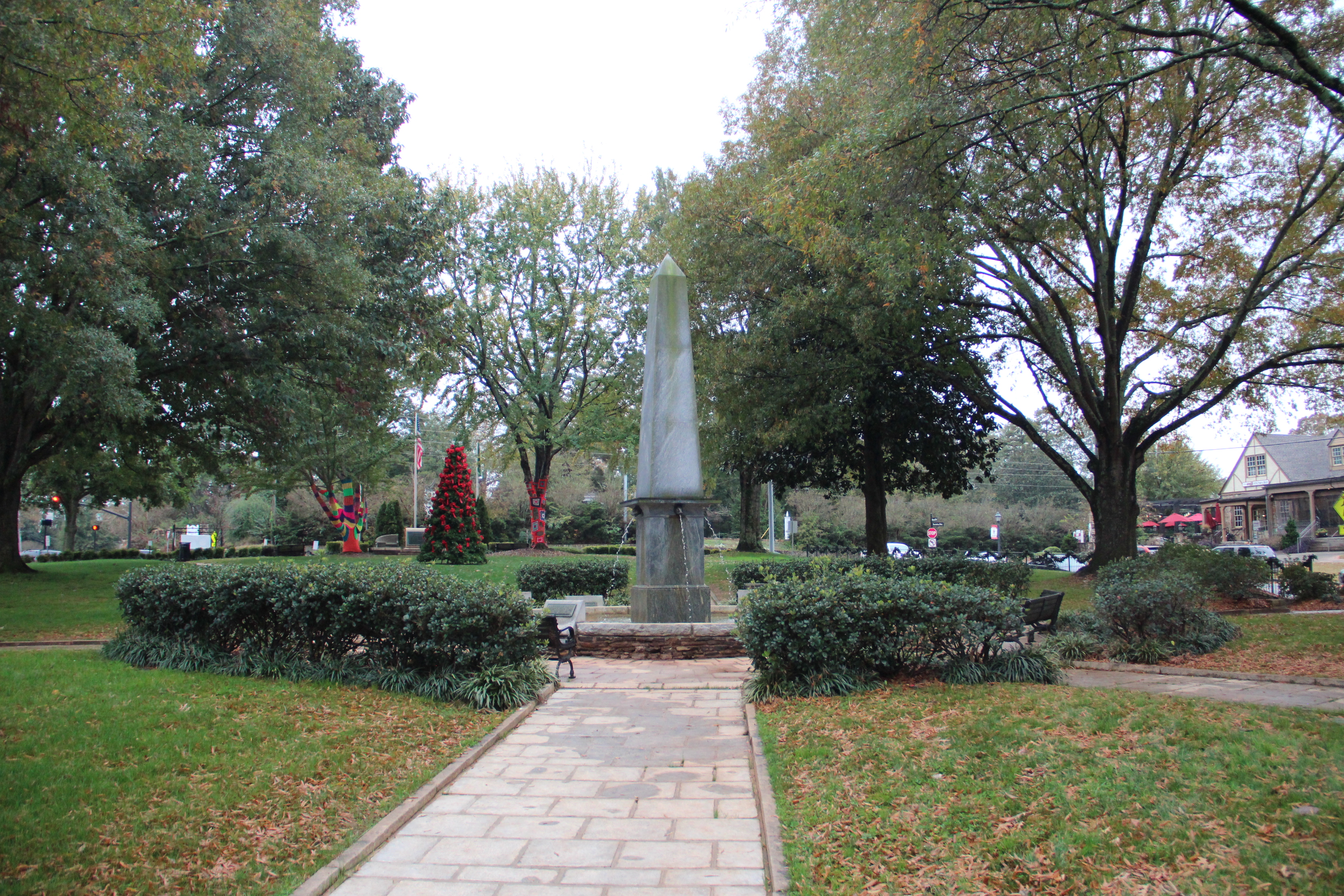 Brick pathway leading to a stone obelisk in a park with large trees and autumn leaves.