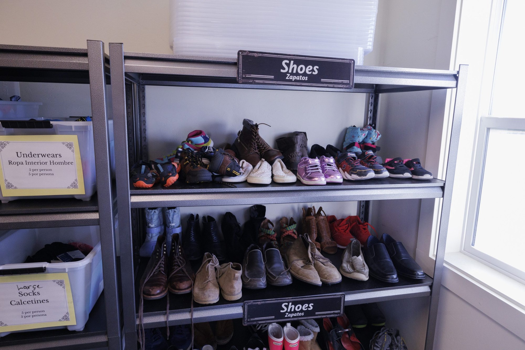 Metal shelves holding donated shoes and boots labeled in English and Spanish.