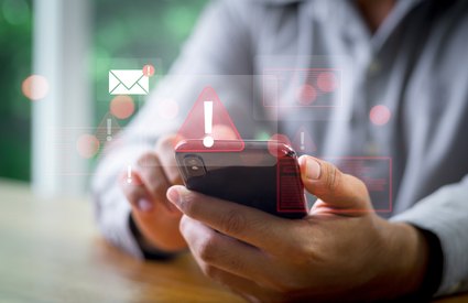 Person in gray hoodie holds a smartphone showing a red warning triangle and email alert icons.