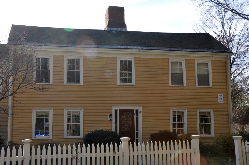A two-story yellow colonial house with white trim, picket fence, and brick chimney.