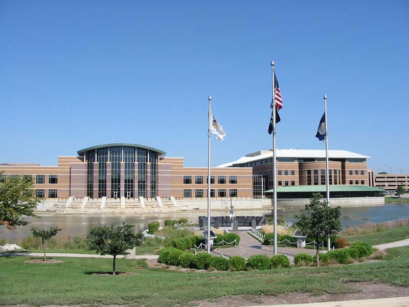 Large brick and glass government building with three flagpoles beside a reflecting pond.