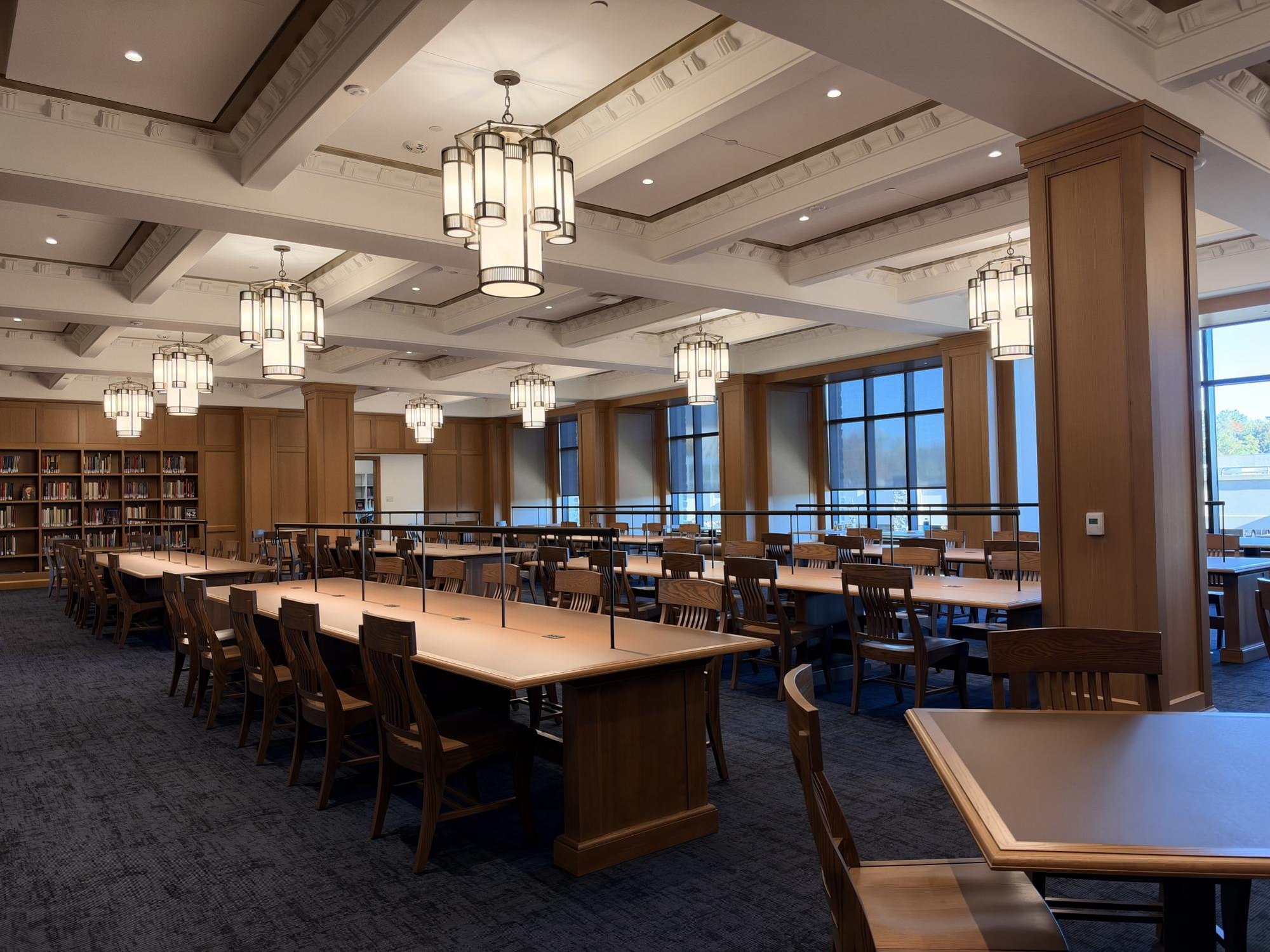 Spacious library reading room with long wooden tables, pendant lights, and tall windows.