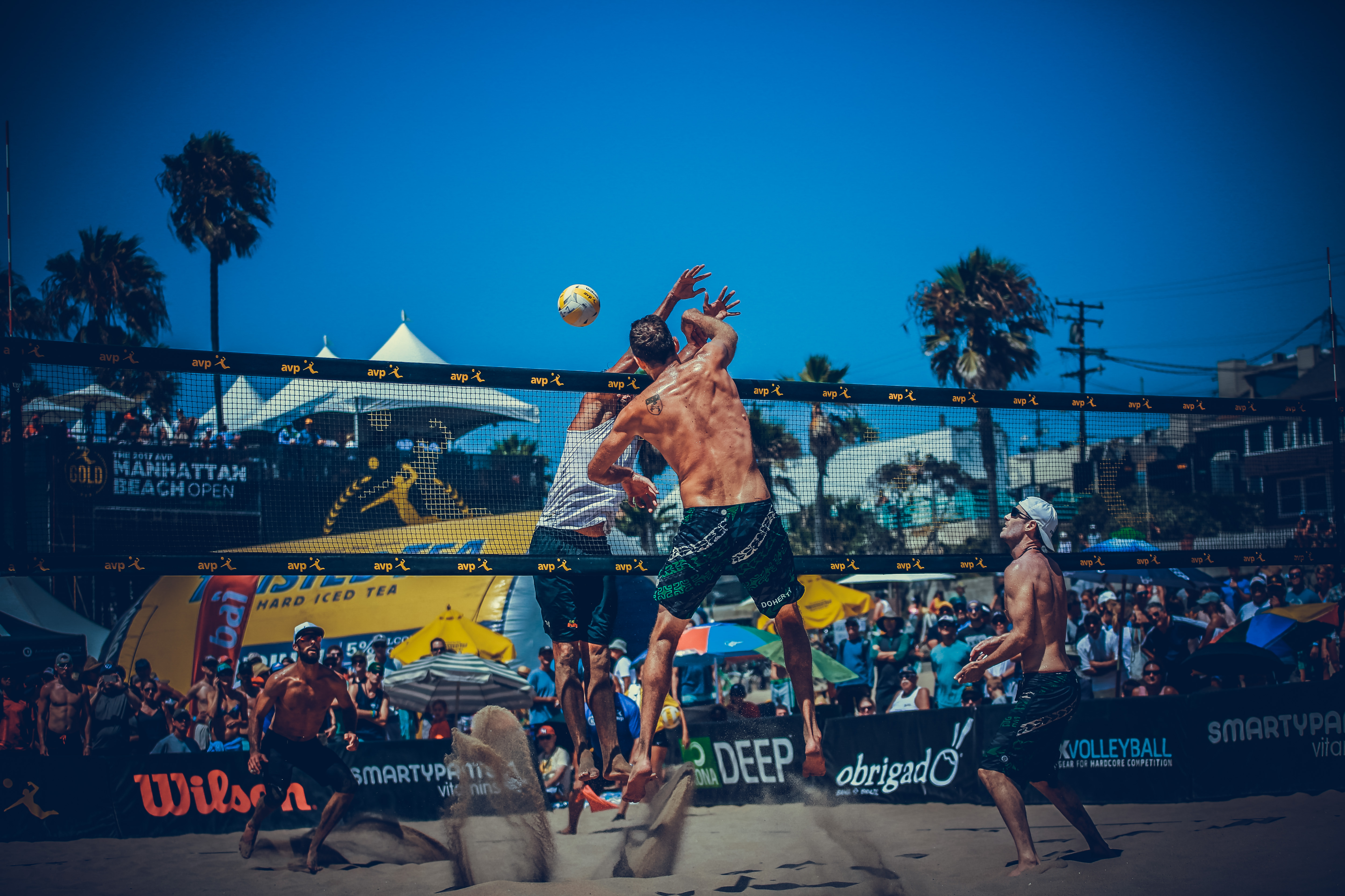 Two beach volleyball players jump at the net with a Manhattan Beach Open banner in the background.