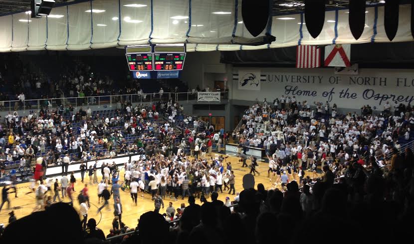 Packed indoor basketball arena with fans on the court and a scoreboard visible overhead.