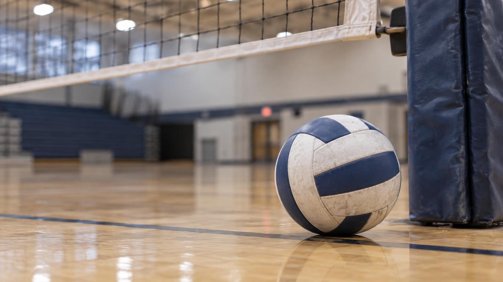 A volleyball resting on a hardwood gymnasium floor near the net post.