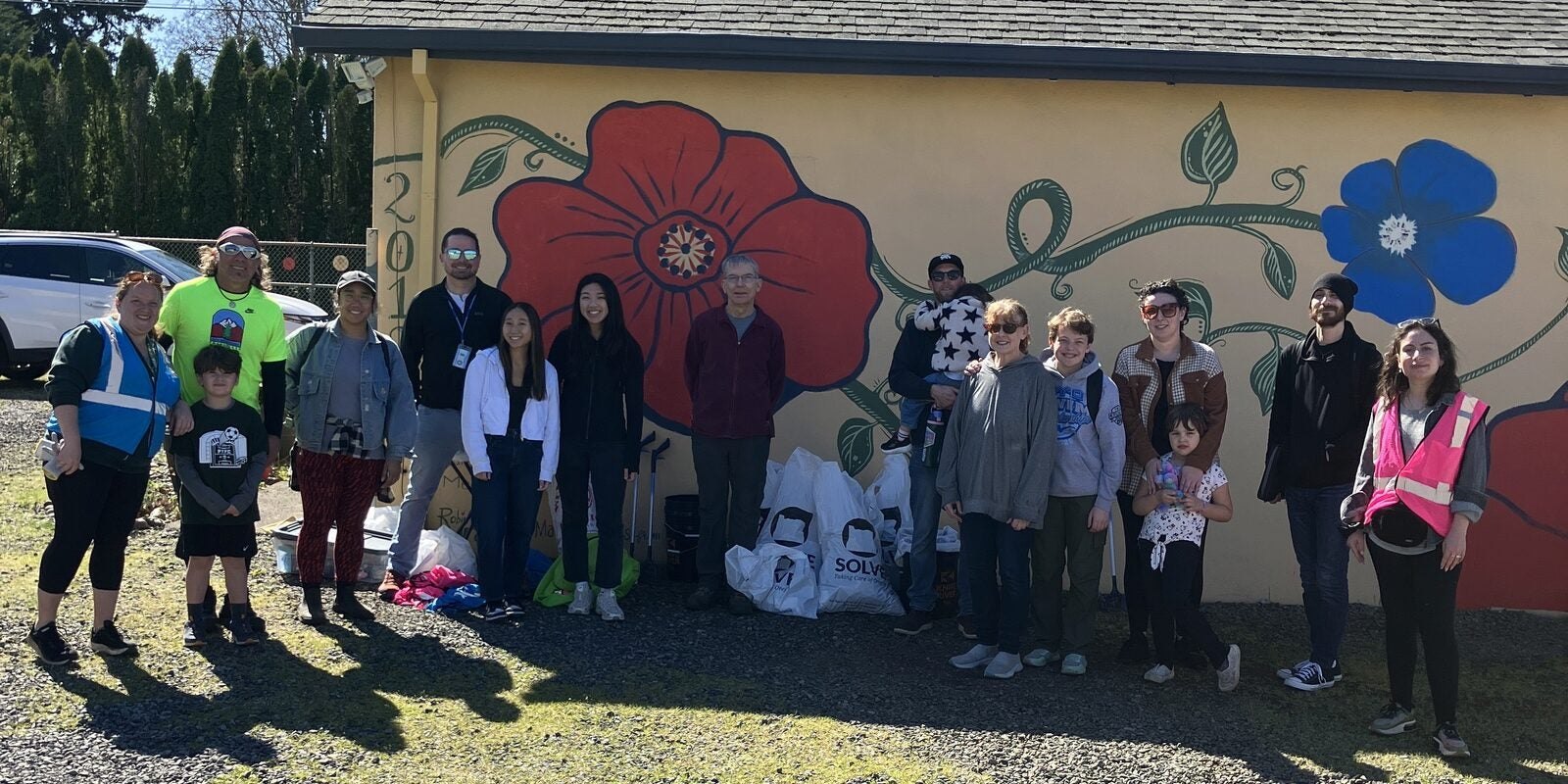 Group of adults and children in safety vests holding trash bags in front of a floral mural.