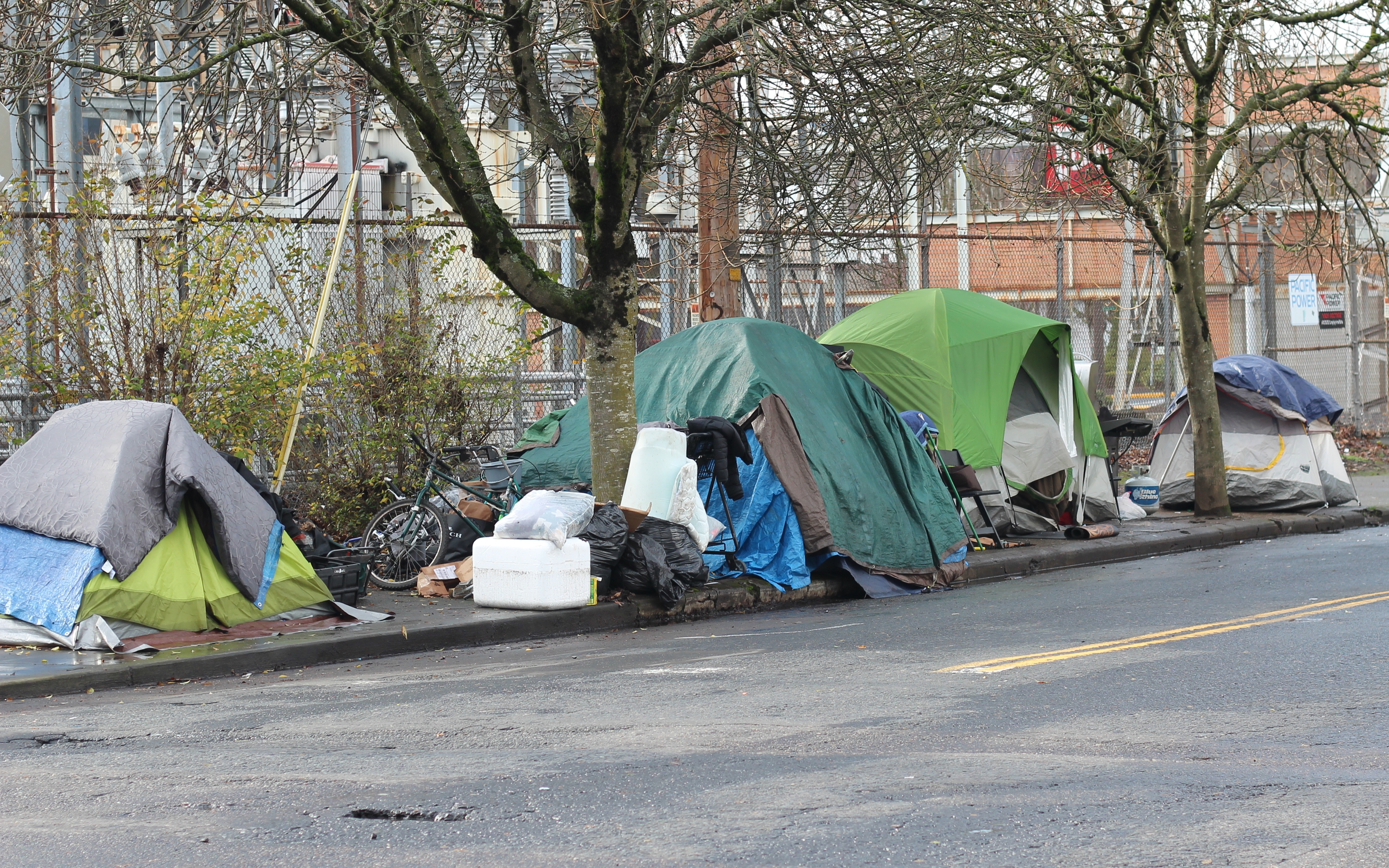 Multiple tents and personal belongings clustered along an urban sidewalk next to a road.