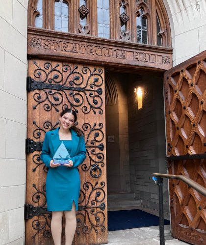 Young woman in a teal outfit holds a triangular trophy outside a Gothic stone archway.