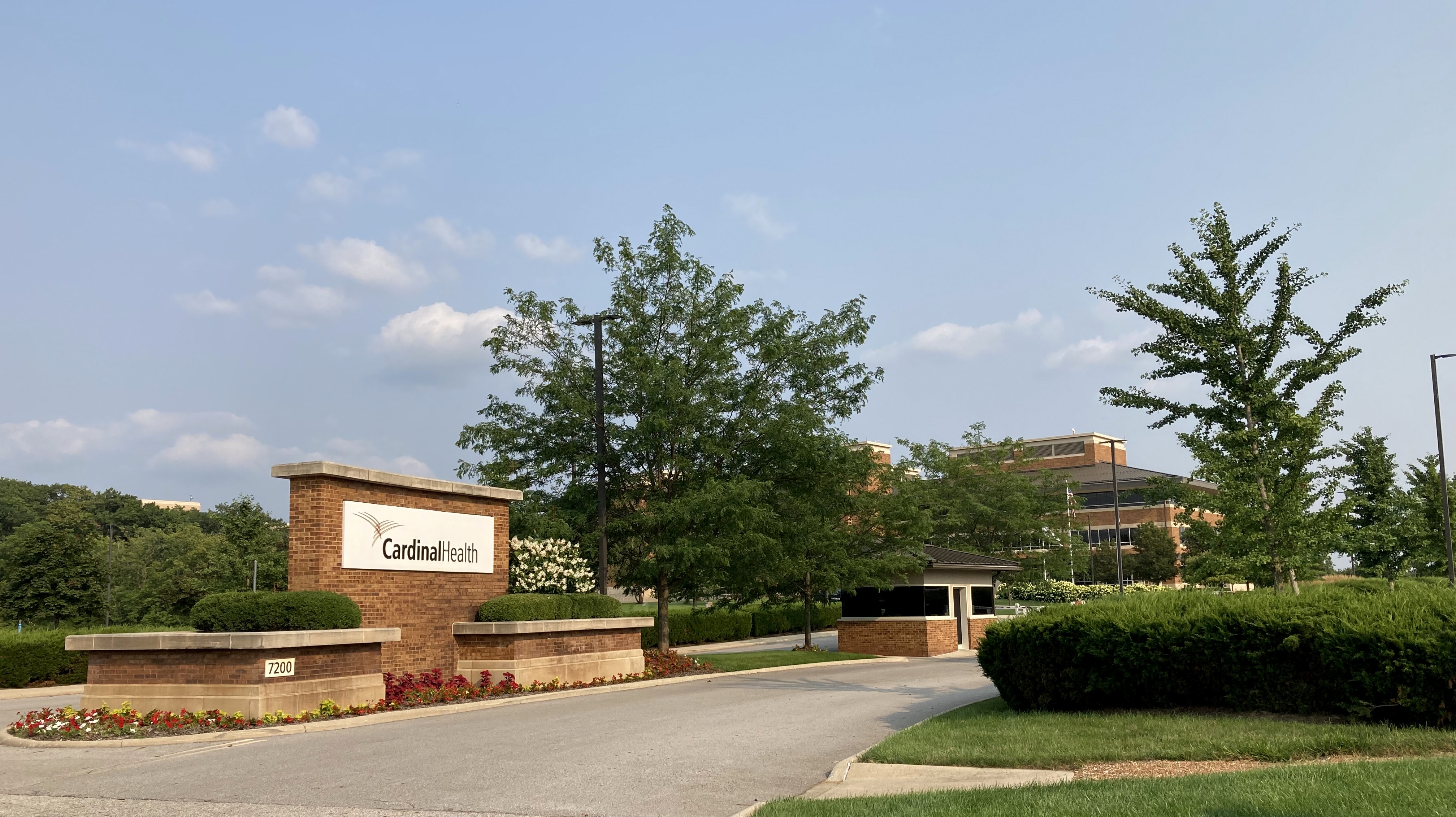 Brick monument sign at Cardinal Health headquarters entrance with office building behind trees.
