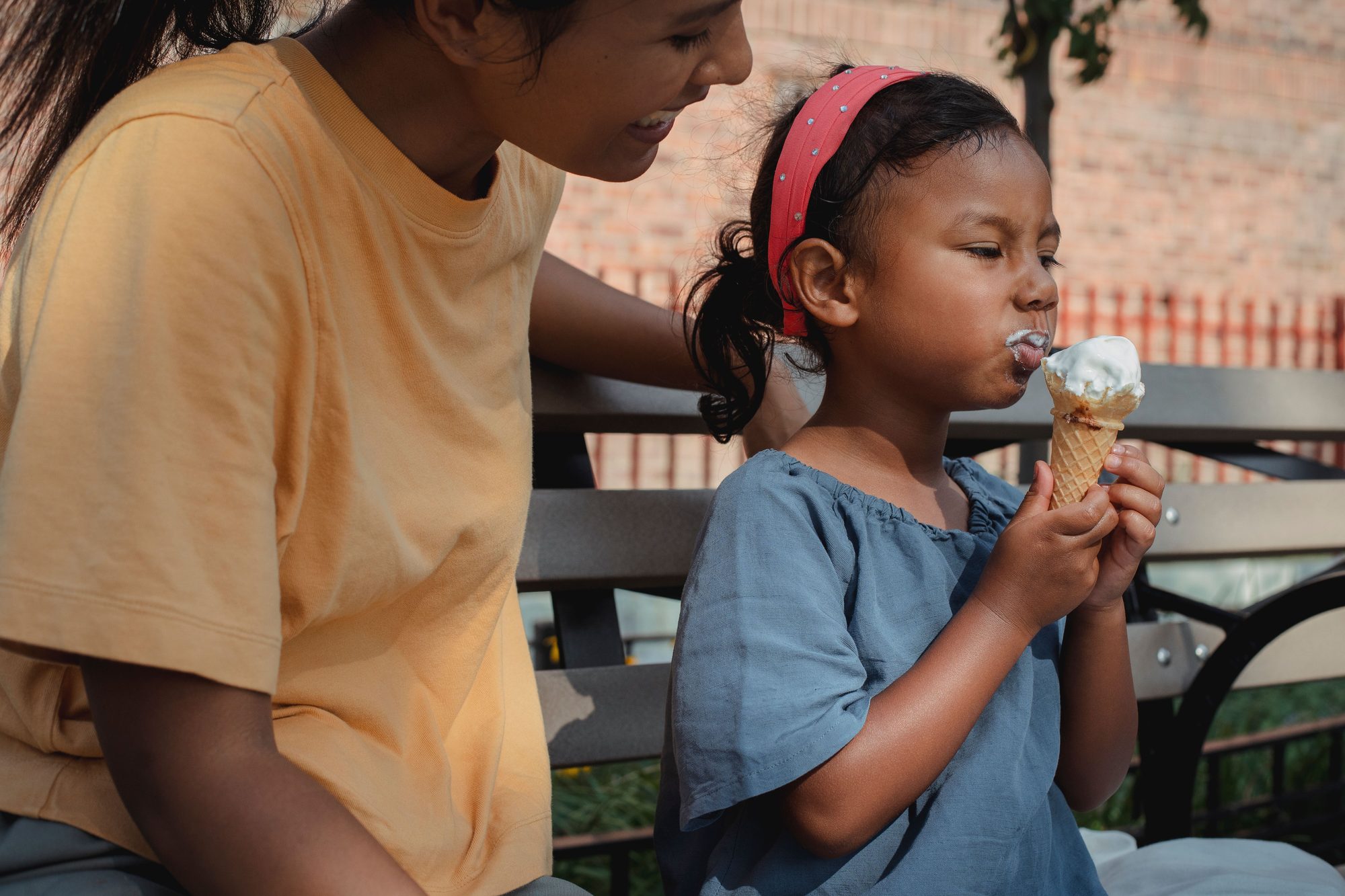 Small Asian girl eating ice cream while sitting on bench with mother on street in sunny day